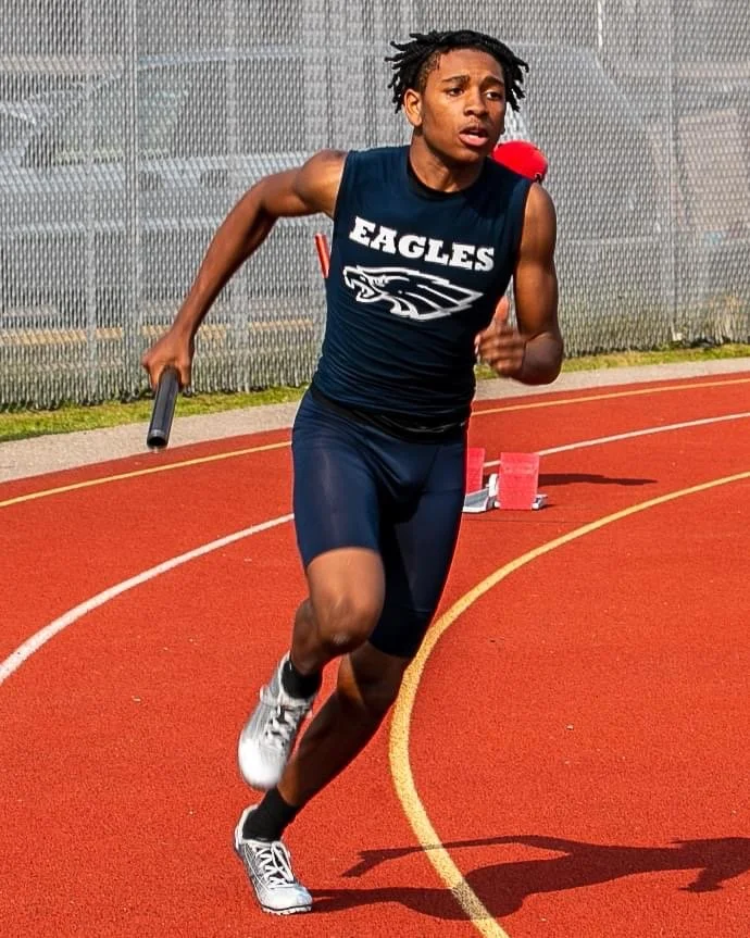 A young male athlete sprints on a red track, holding a relay baton in his right hand, wearing a navy athletic uniform with 'EAGLES' written on the front, and has dreadlocked hair.