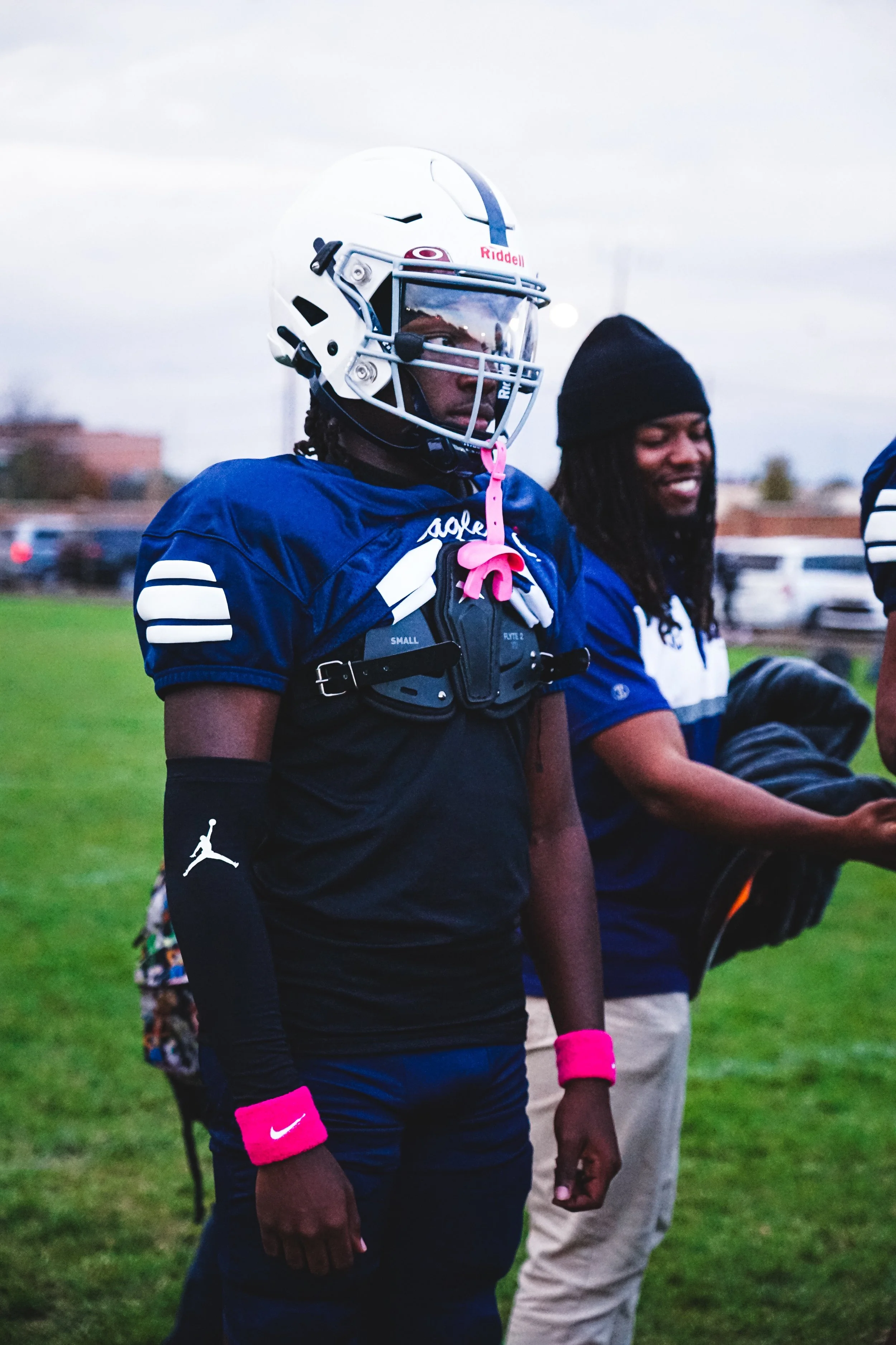 A football player stands on the field wearing a helmet, pads, and game uniform with pink accessories, with another person in casual clothes beside him.