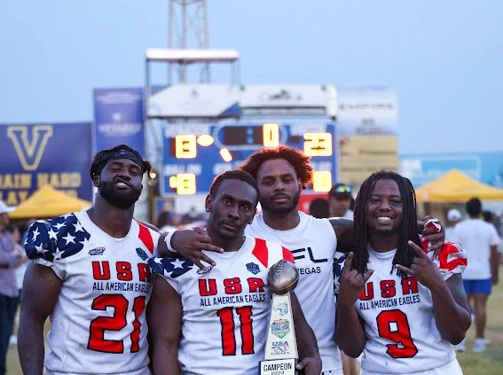 Four young men wearing USA football jerseys posing together at a sports event with a scoreboard in the background.