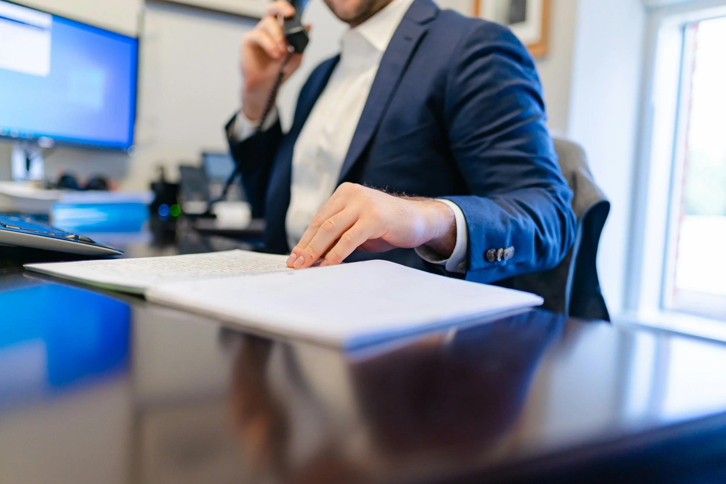 A man in a business suit sitting at a desk, talking on a landline phone, with a notebook and office equipment around him.
