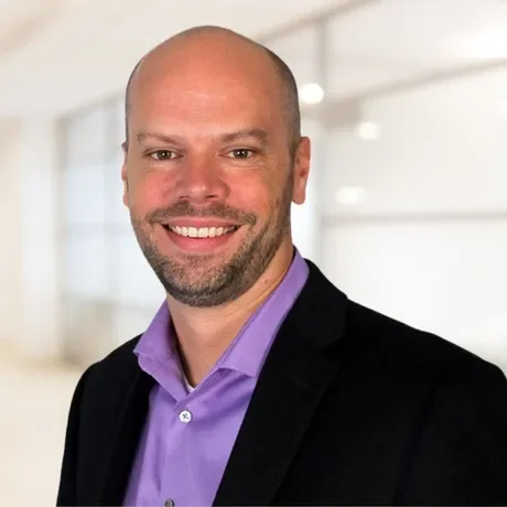 Portrait of a smiling man with a bald head and beard, wearing a purple dress shirt and black blazer, in a modern indoor setting.