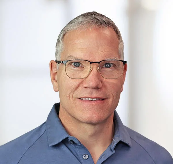 Headshot of a middle-aged man with short gray hair, wearing glasses and a blue collared shirt, smiling at the camera.