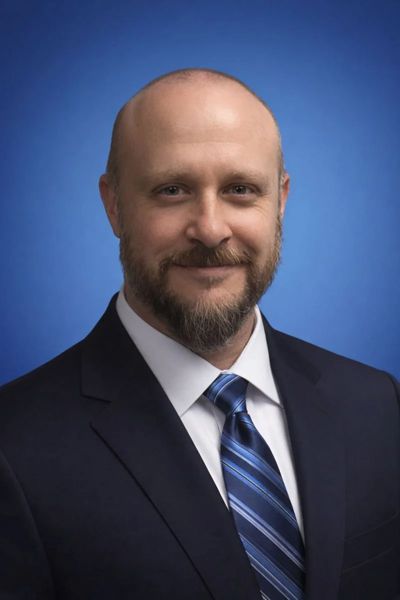 Professional headshot of a man with a beard, wearing a navy suit, white shirt, and striped blue tie, against a blue background.
