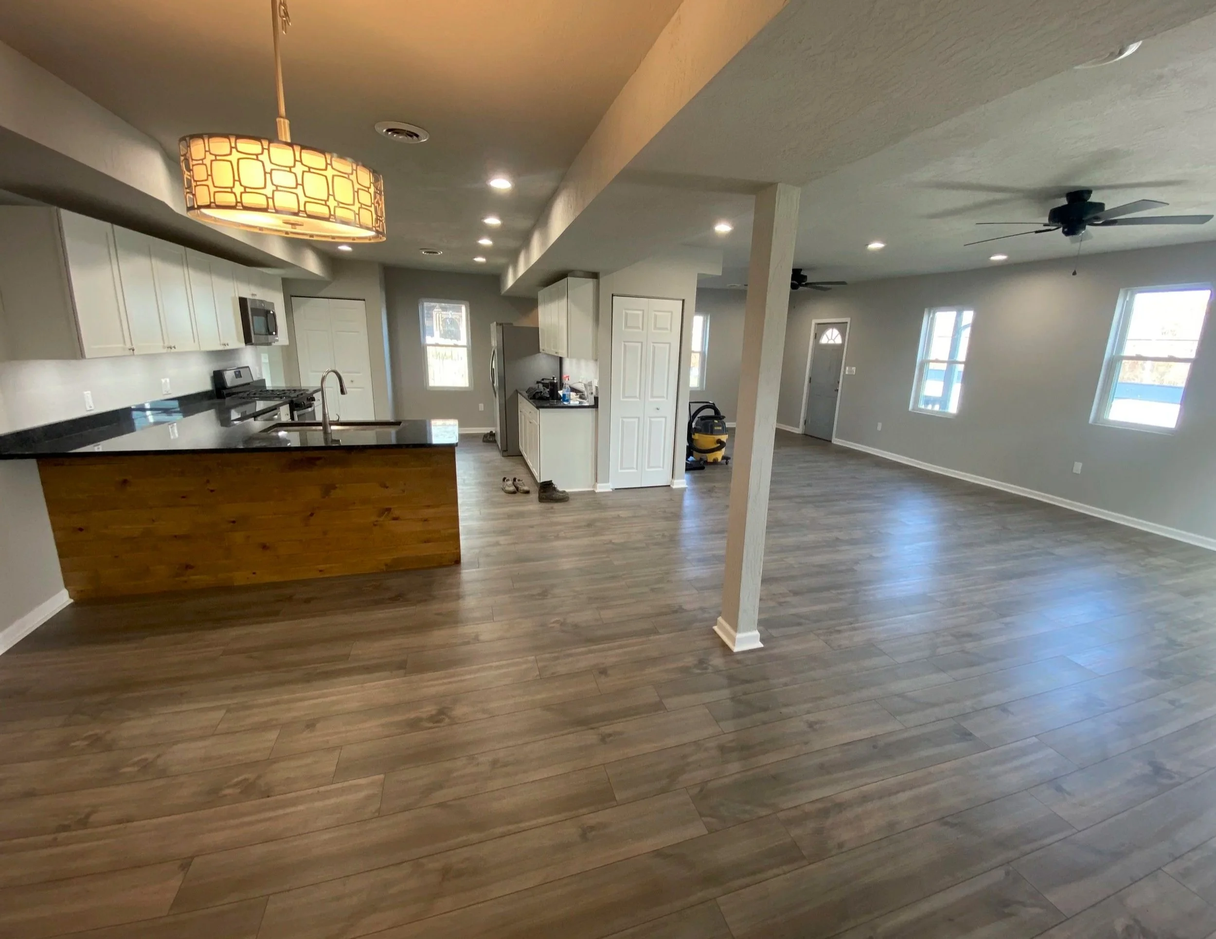 Empty open-concept living space with a kitchen, hardwood floors, and windows, with ceiling fans and a vacuum in the background.
