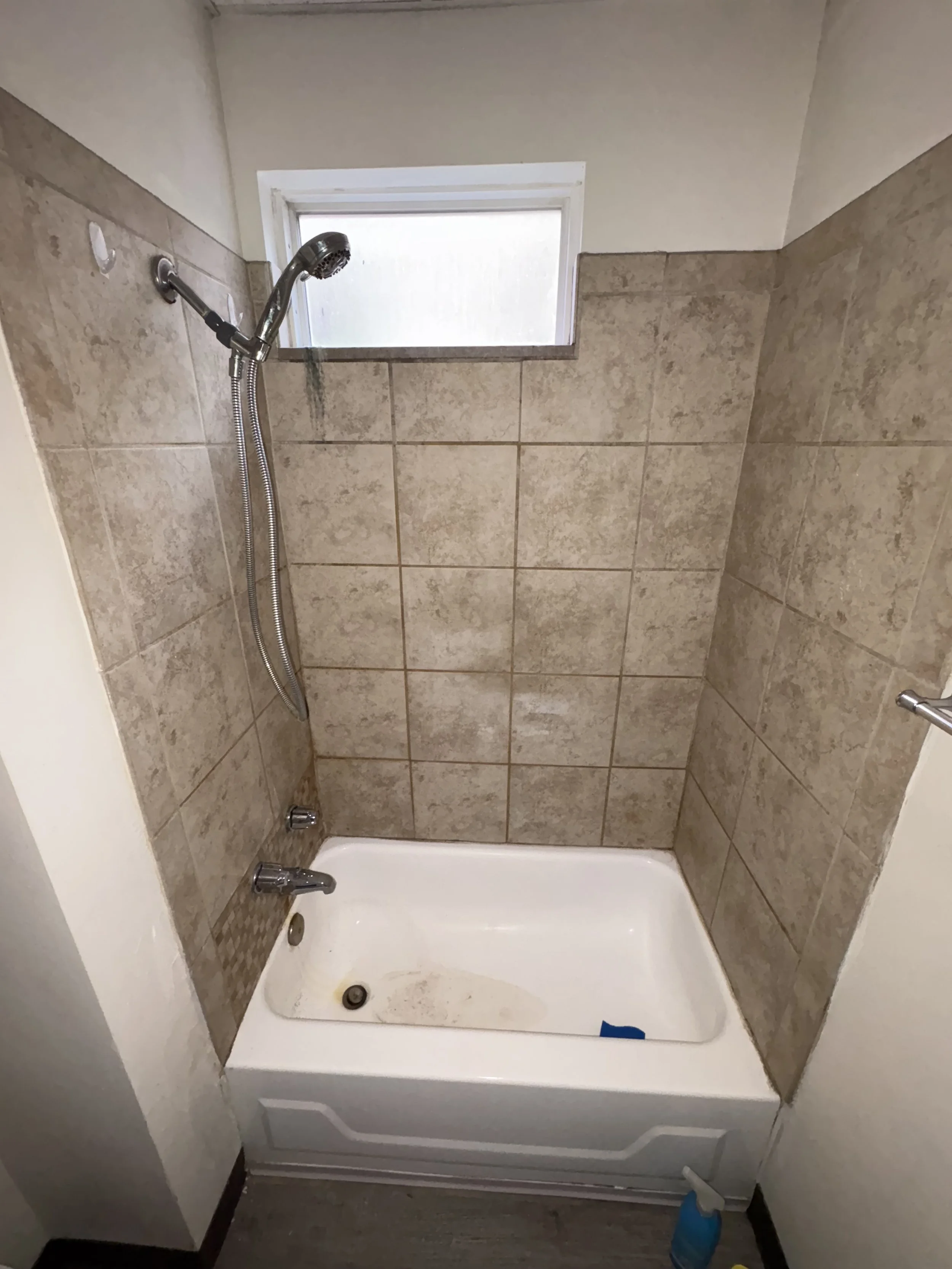 A small bathtub with beige tile surround and a sliding window above. The bathtub appears to be dirty with a soap dish, a blue cleaning product, and a wall-mounted handheld showerhead.