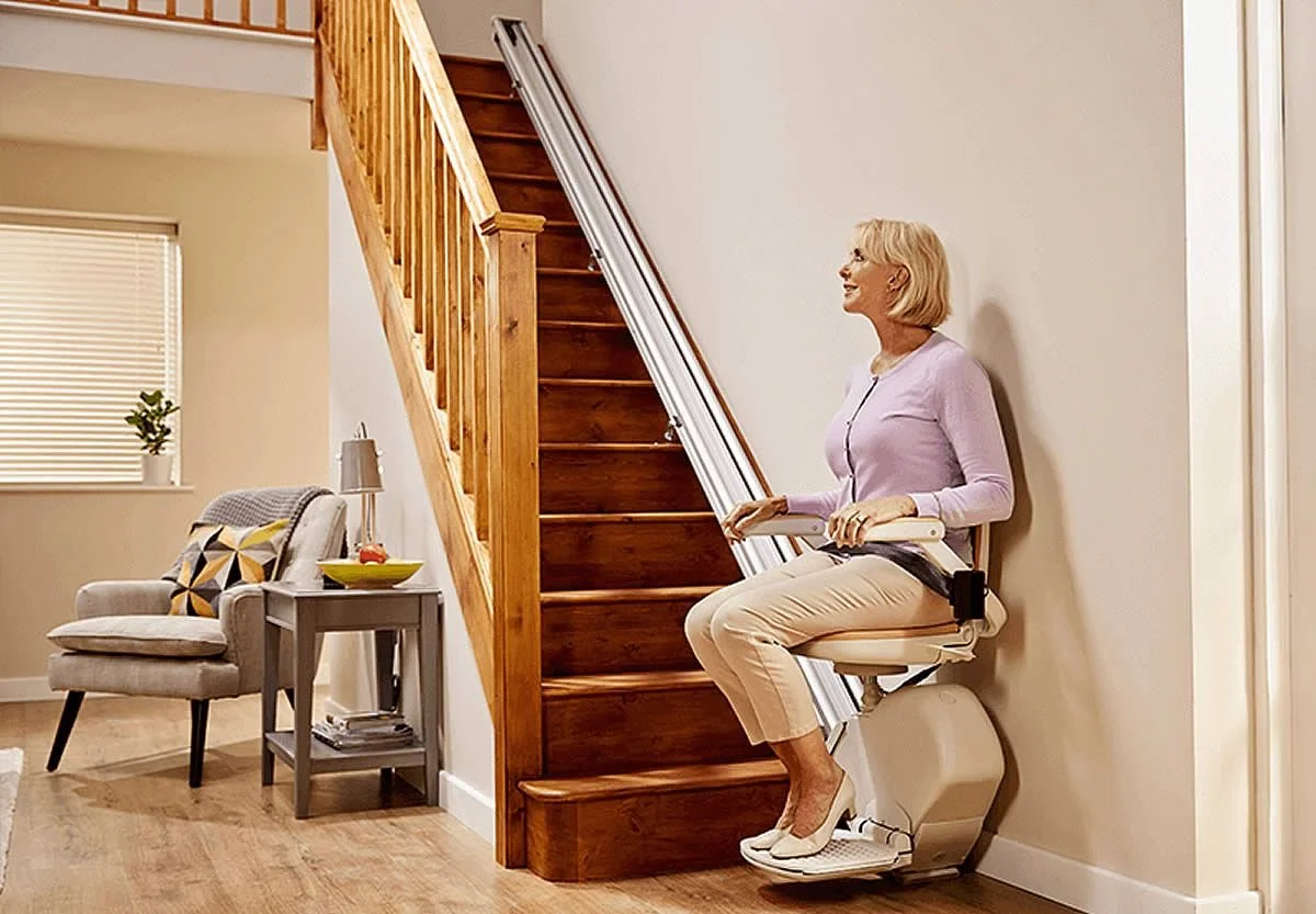 A senior woman using a stair climbing assistive device in her home, sitting on a chair-like stepper near a wooden staircase in a living room.