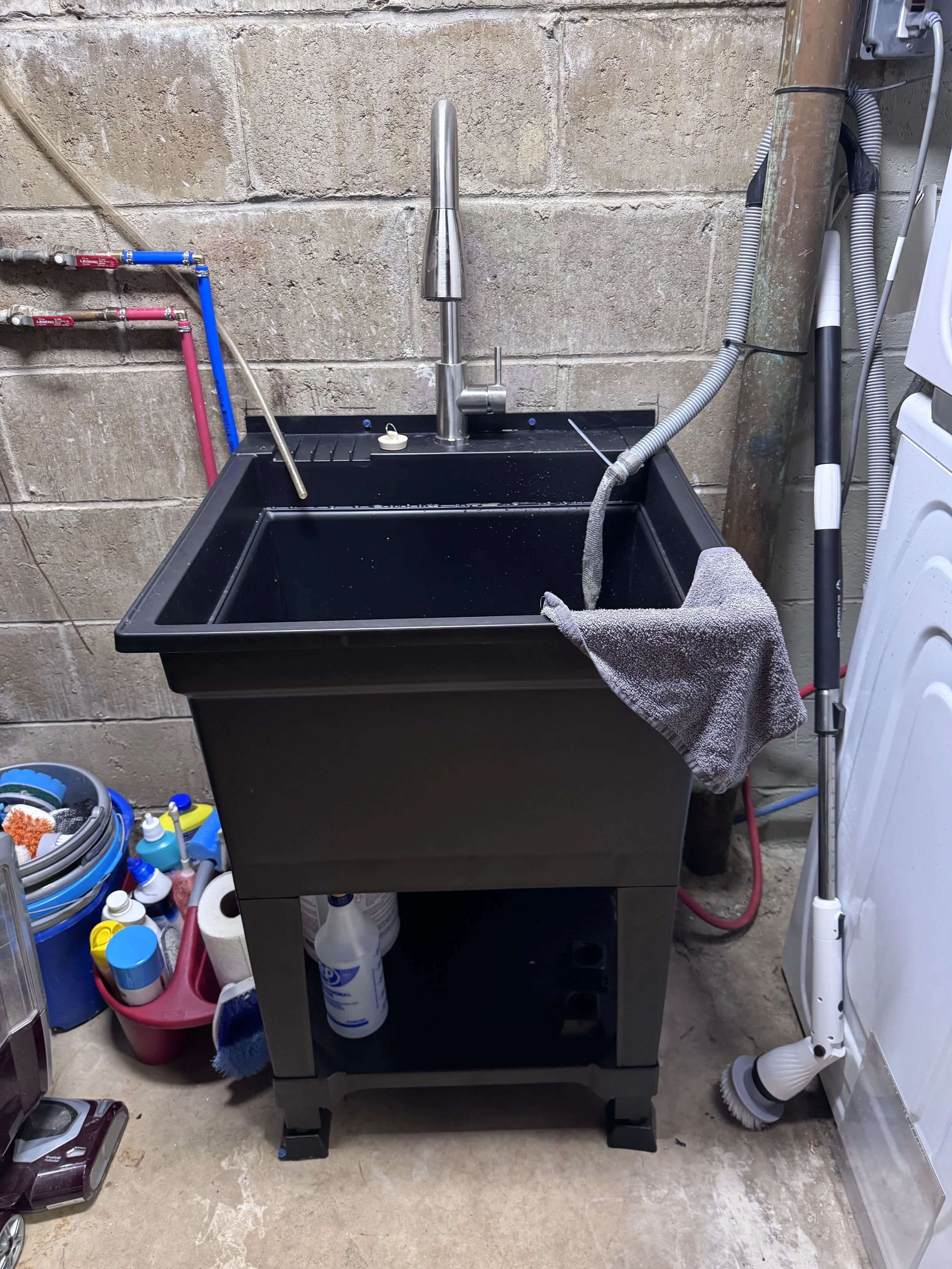Black utility sink with cleaning supplies underneath, a gray cloth hanging on the faucet, and laundry attachments nearby in a laundry or utility room.