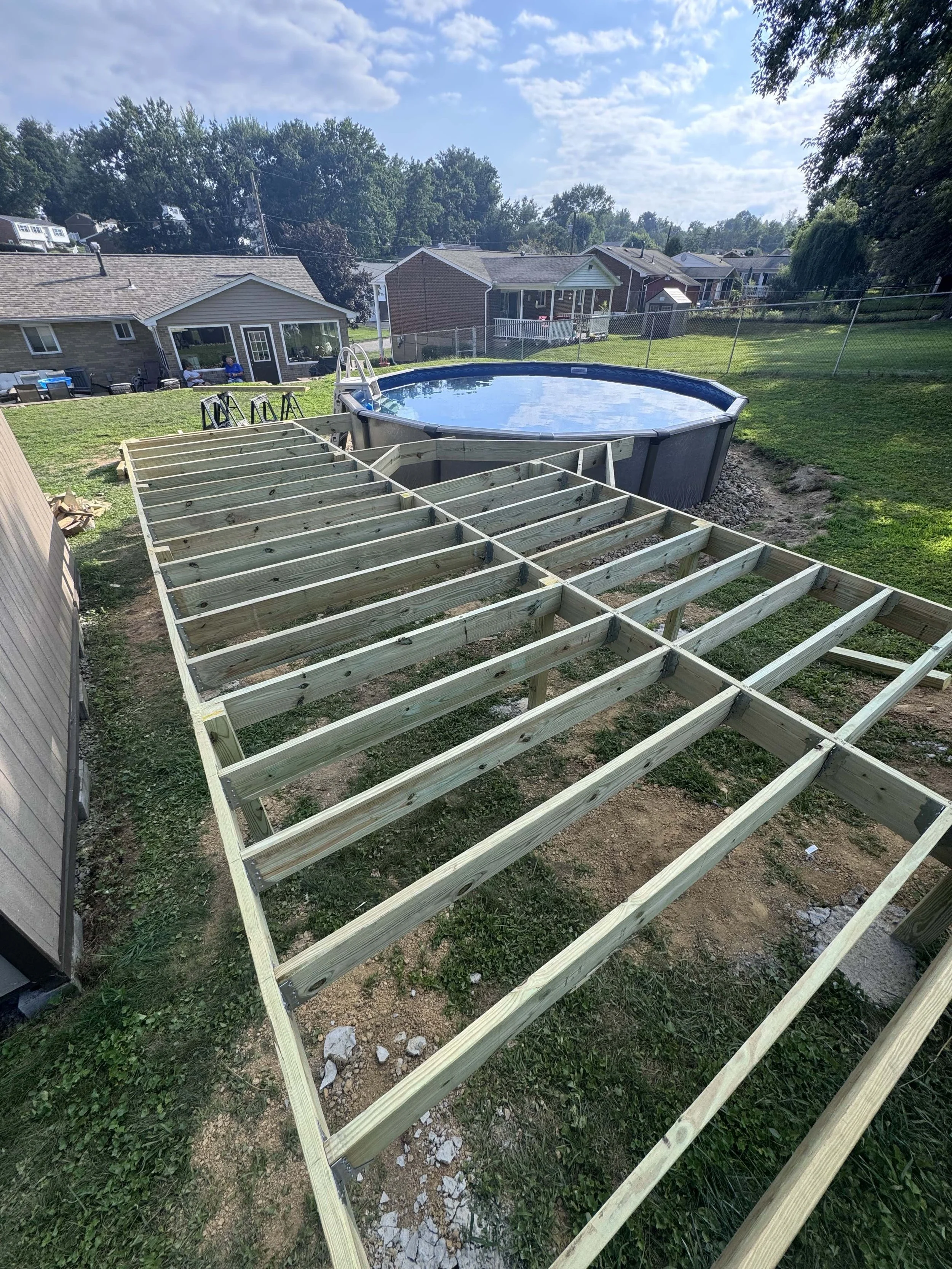 Wooden deck platform under construction in backyard with above ground pool, surrounded by grass, neighboring houses, and trees in the background.