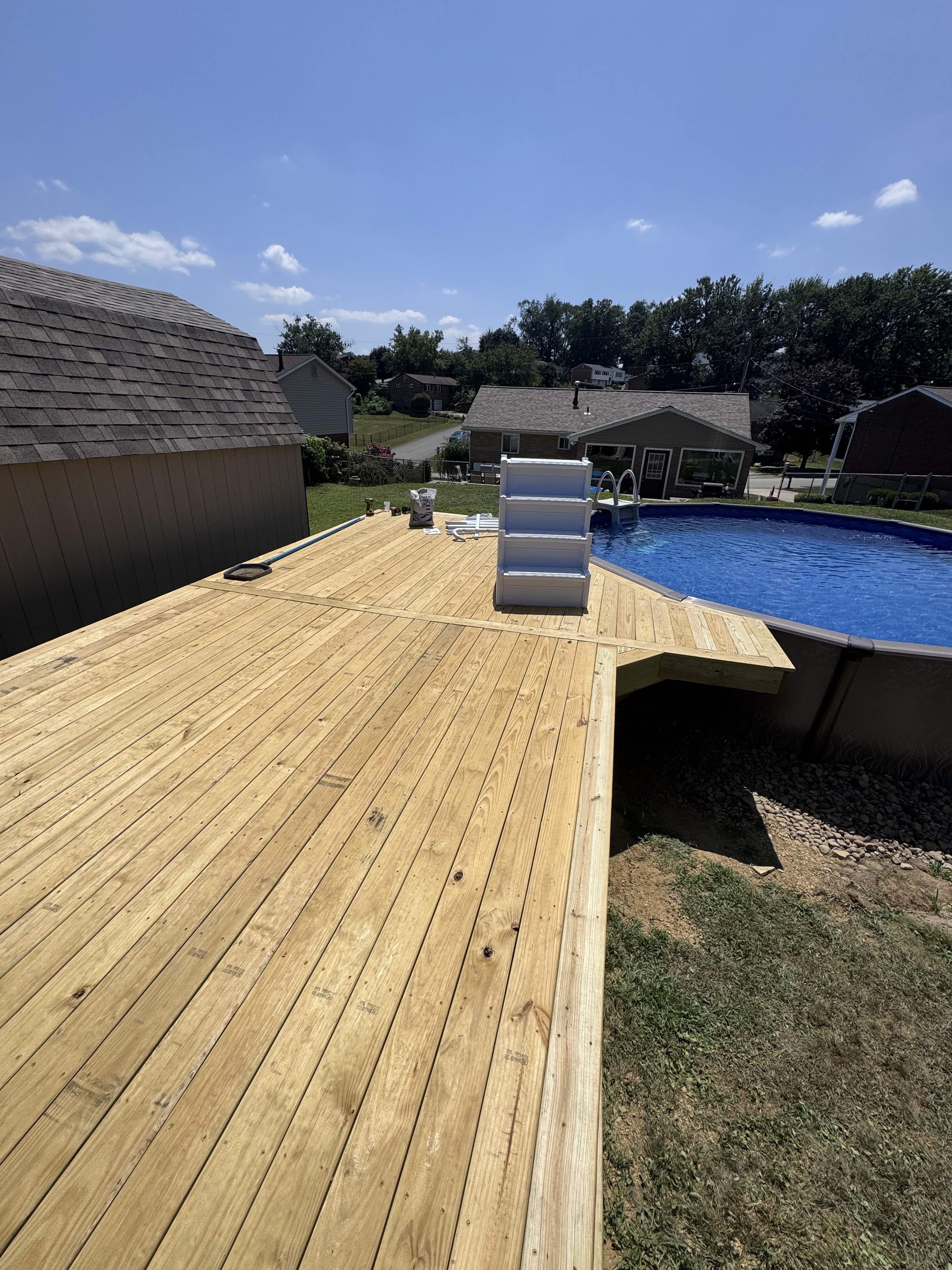 A newly built wooden deck surrounding an above-ground swimming pool in a backyard under a blue sky with clouds.