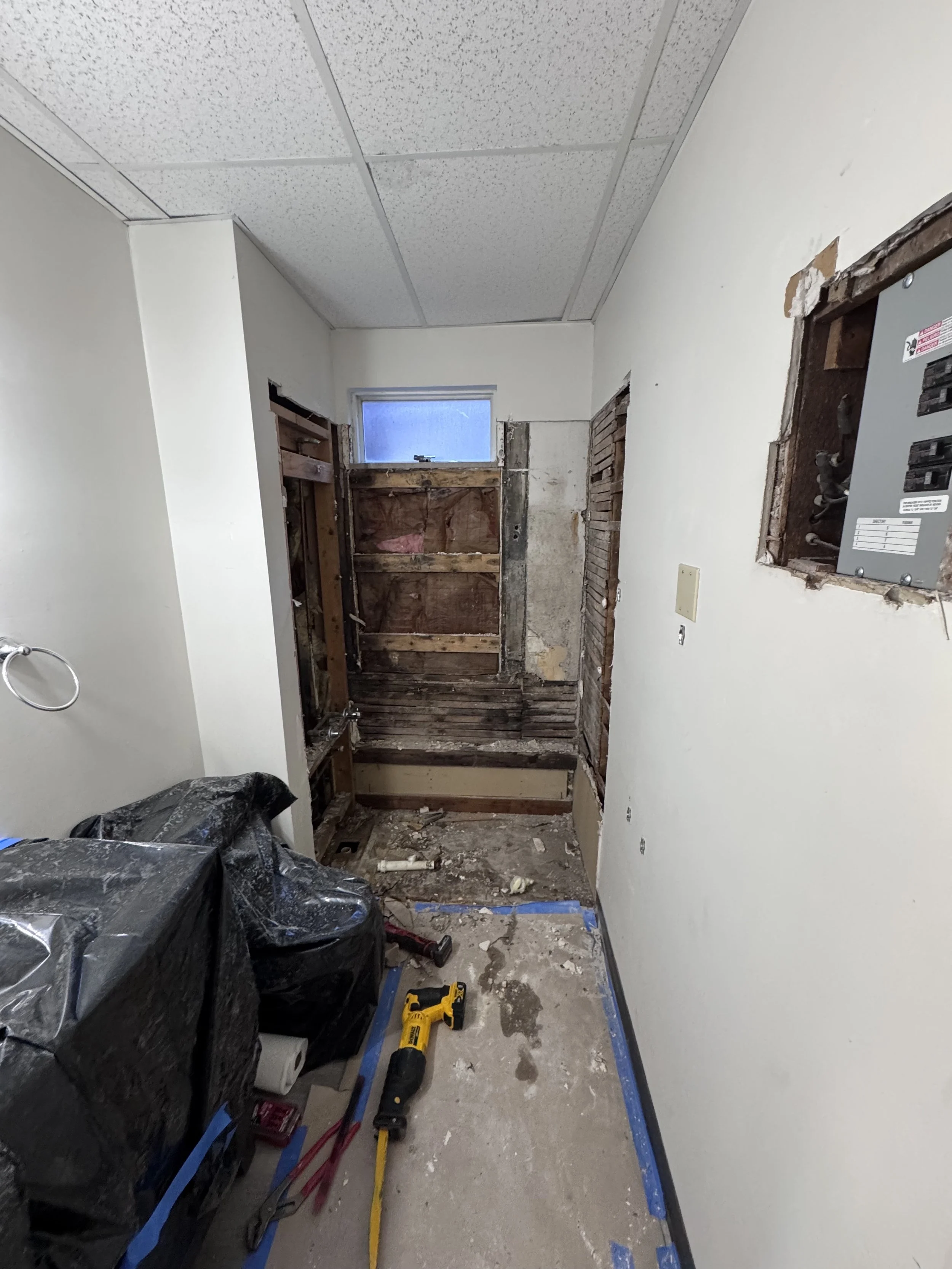 Bathroom under renovation with exposed plumbing and electrical panel, construction tools, and debris on the floor.