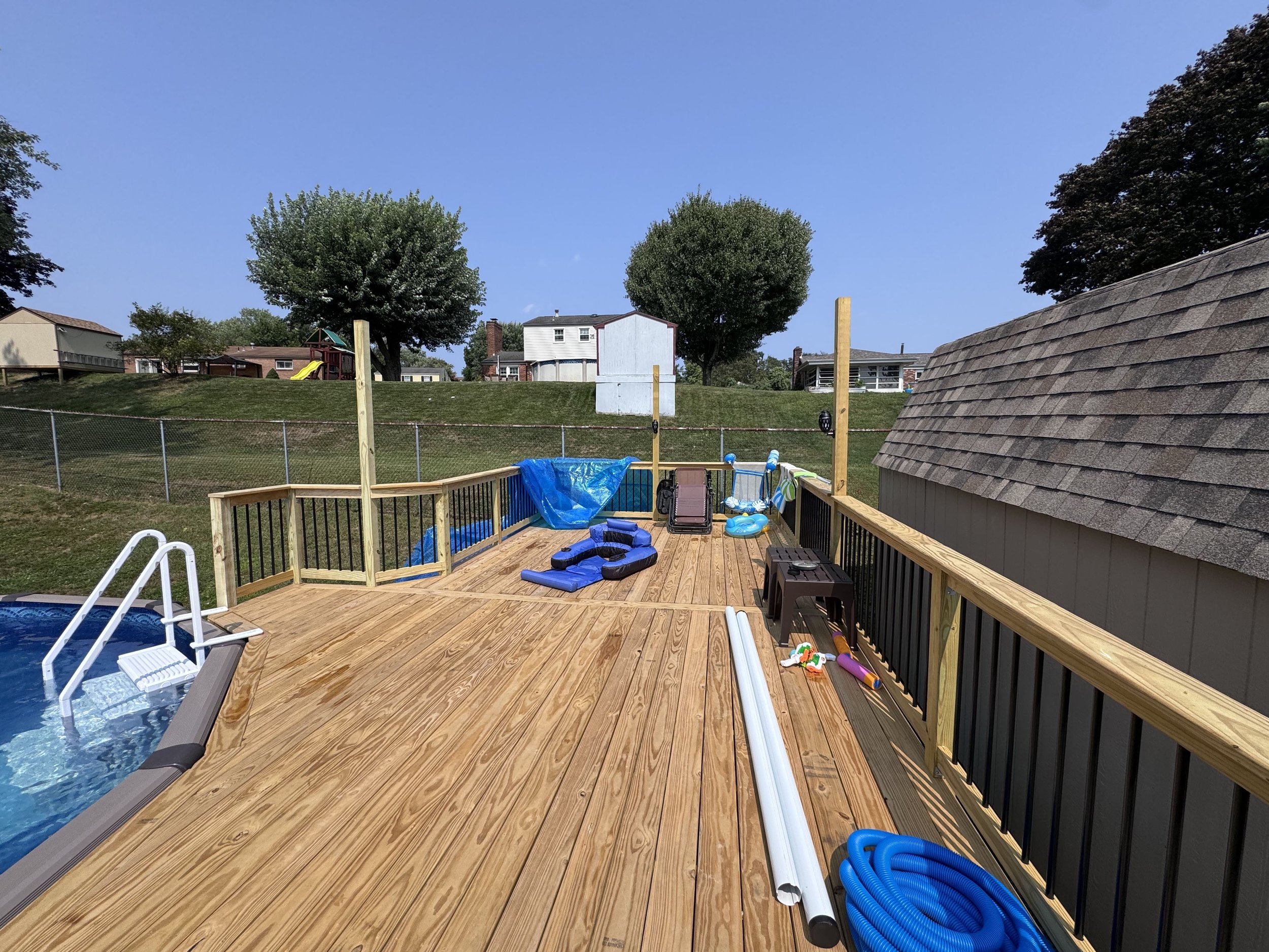 A newly built wooden deck with a pool, pool toys, and construction materials in a backyard with a grassy hill, trees, and neighboring houses.