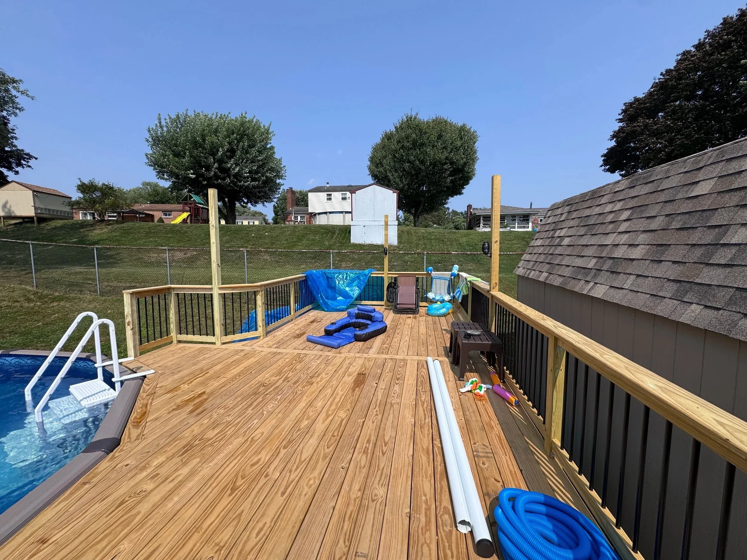 Backyard deck with pool and children's toys, including inflatable floaties, pool noodles, and a small slide, surrounded by a fence, with neighboring houses and trees in the background under a clear blue sky.