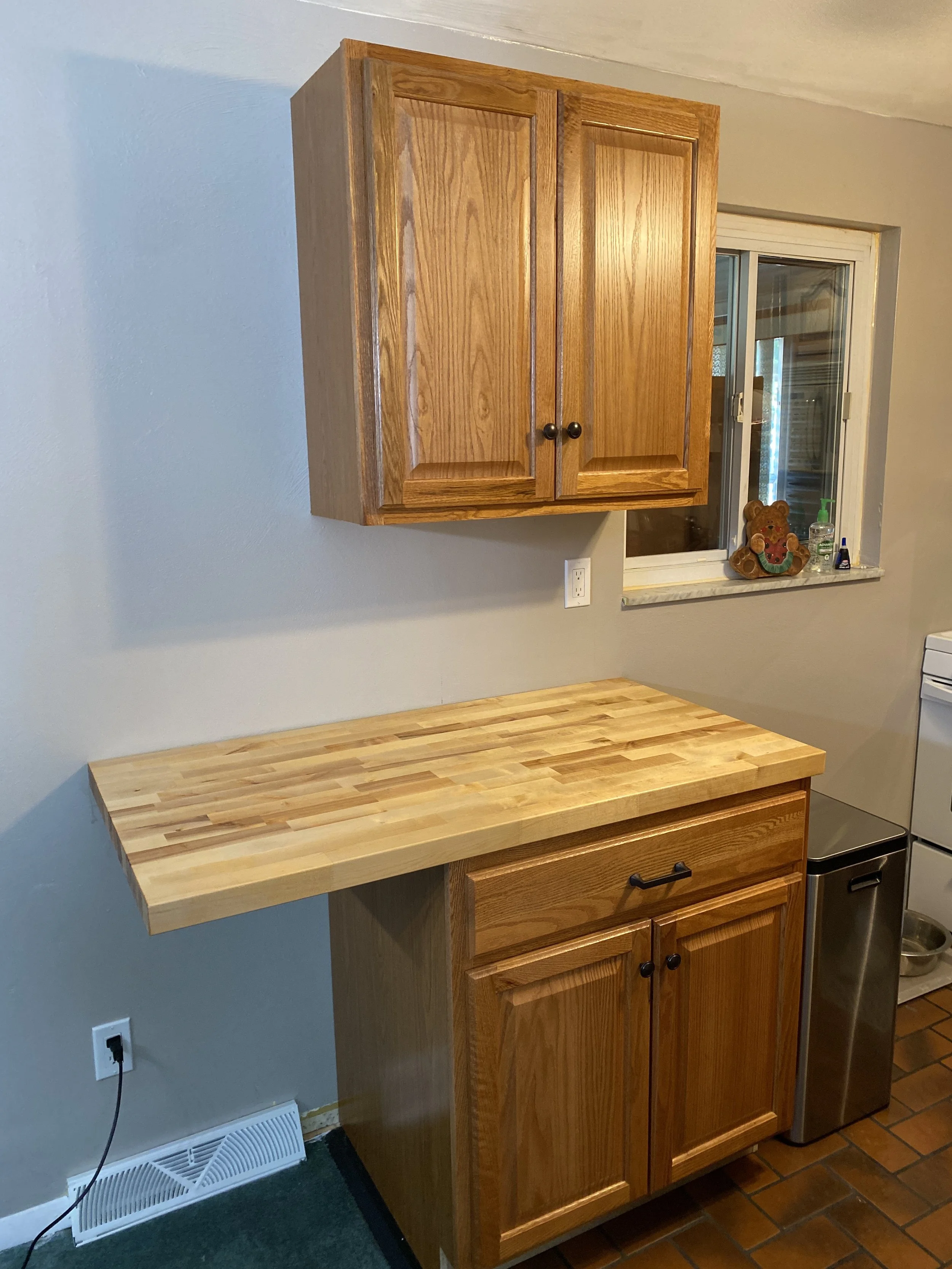 Kitchen with wooden cabinets and a butcher block countertop, window with decorative teddy bear, and small appliances.