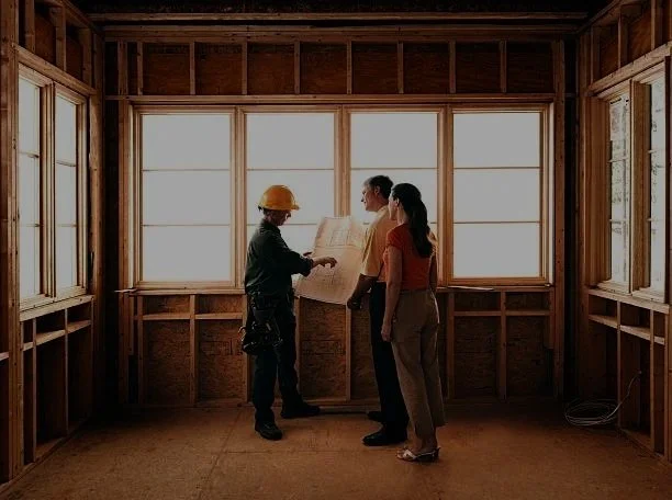 A construction worker in a hard hat consulting with a couple inside an unfinished house with exposed wooden framing and large windows.