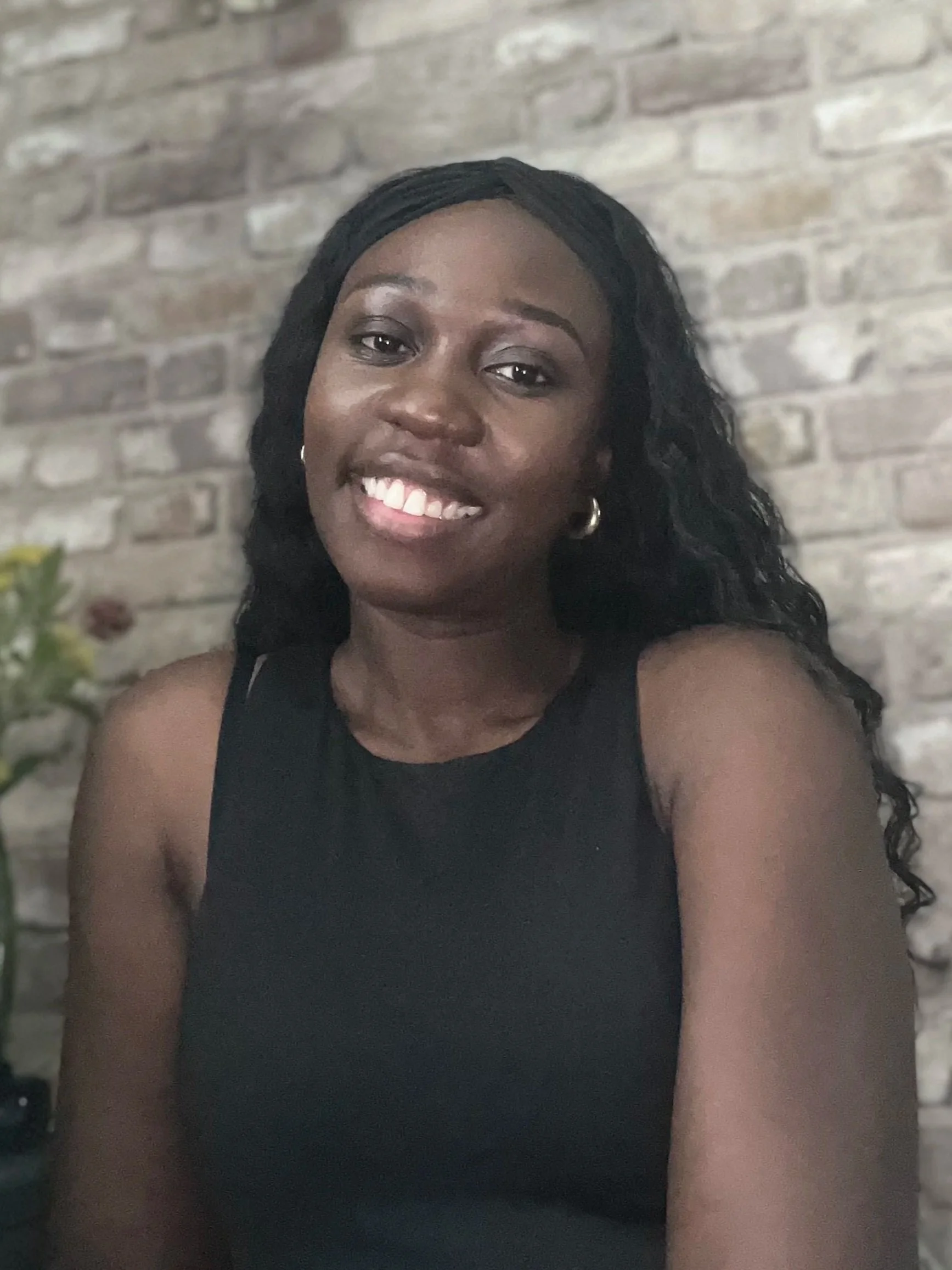 Dr Alria Williams smiling with long curly black hair, wearing a sleeveless black top, sitting in front of a brick wall.