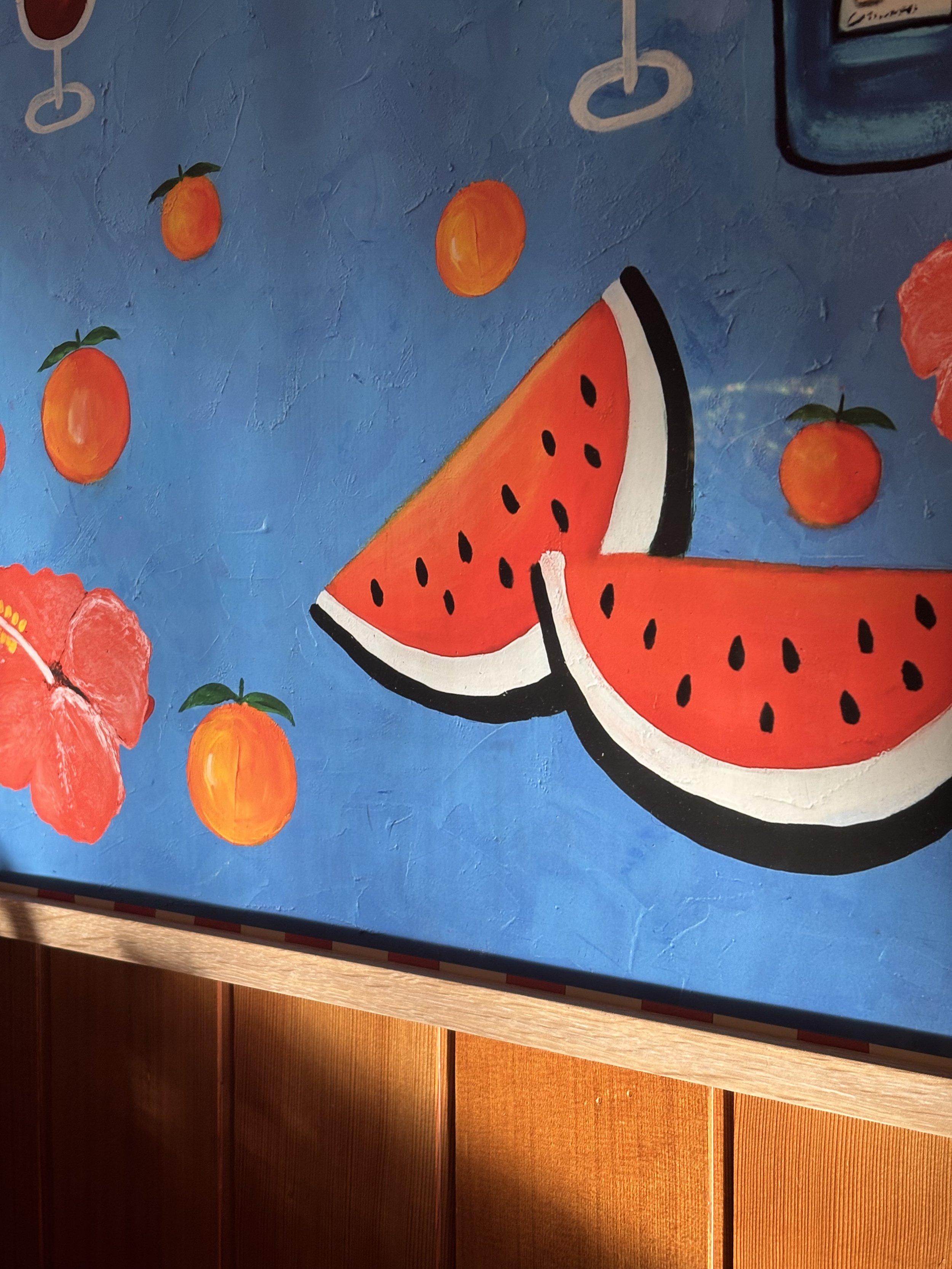 Hand-painted mural featuring a large slice of watermelon with black seeds on a blue background, surrounded by small orange fruit illustrations with green leaves and hanging rings.