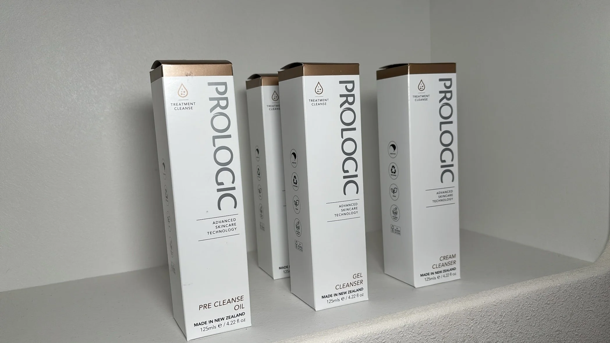Four white boxes of skincare products, labeled 'PROLICIOUS,' arranged on a white shelf against a gray wall. Each box has a bronze-colored top and features icons and text indicating different skincare functions such as pre-cleanse oil, gel cleanser, and cream cleanser. The labels mention advanced skincare technology and that the products are made in New Zealand, with each box containing 125ml or 4.22 fl oz.