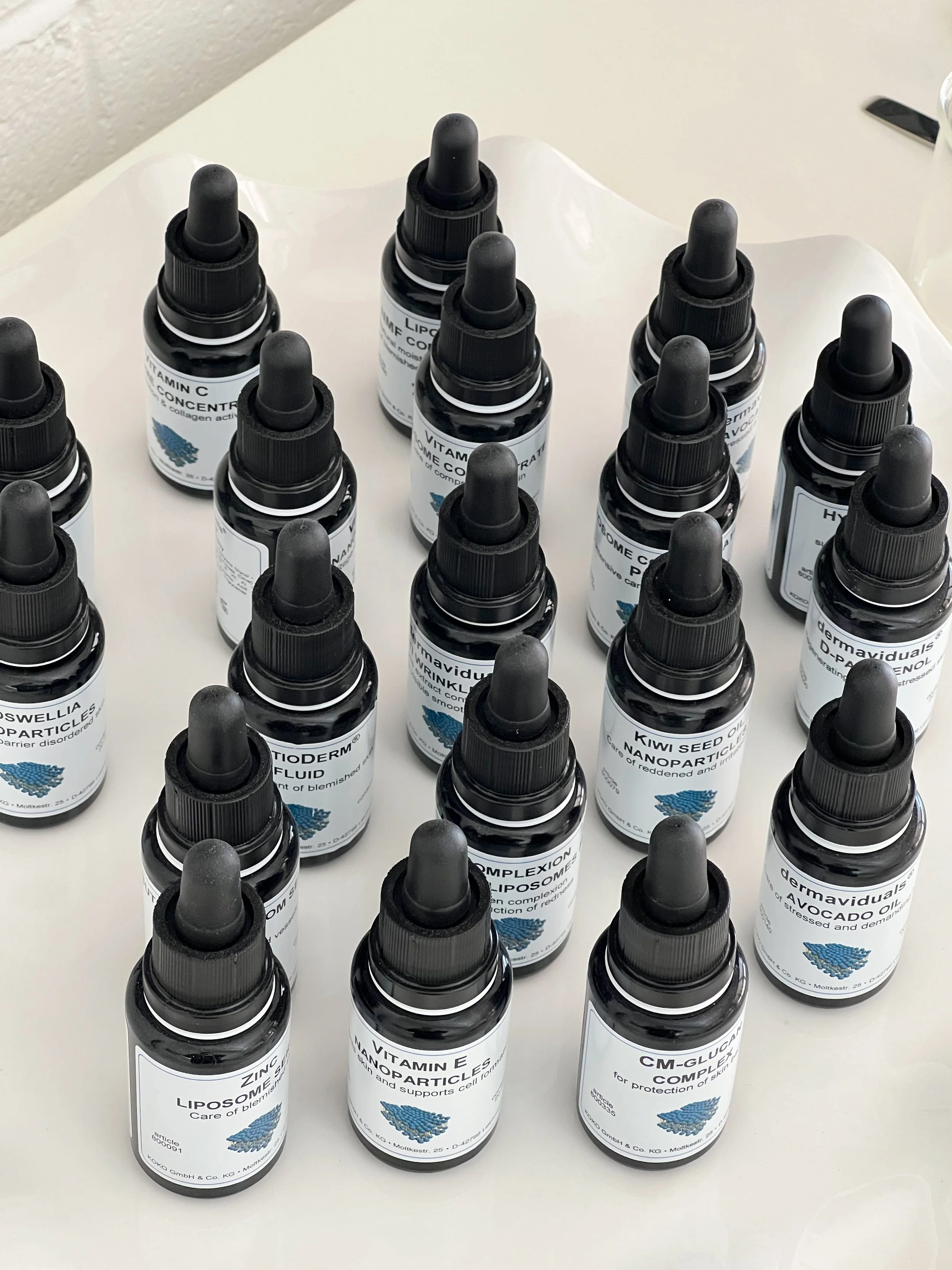 Multiple small dark bottles with droppers on a white surface, labeled with various vitamins and supplements.