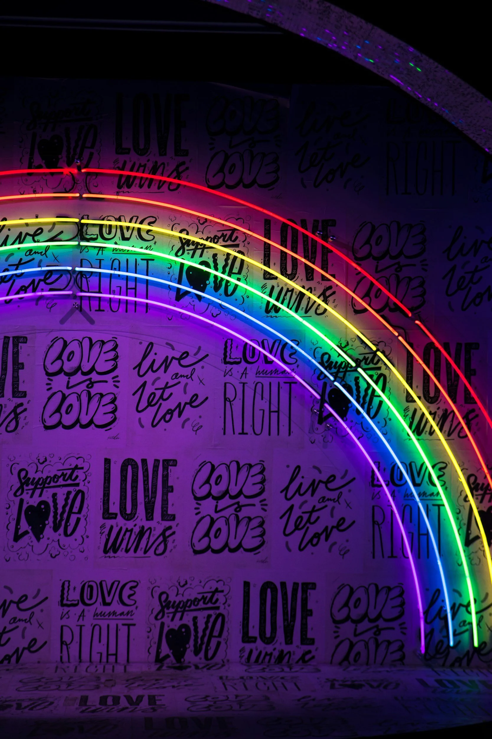 Colorful neon rainbow lights against a background with various love-themed sayings such as 'Love is a human right,' 'Support love,' and 'Live and let love' written in different styles.