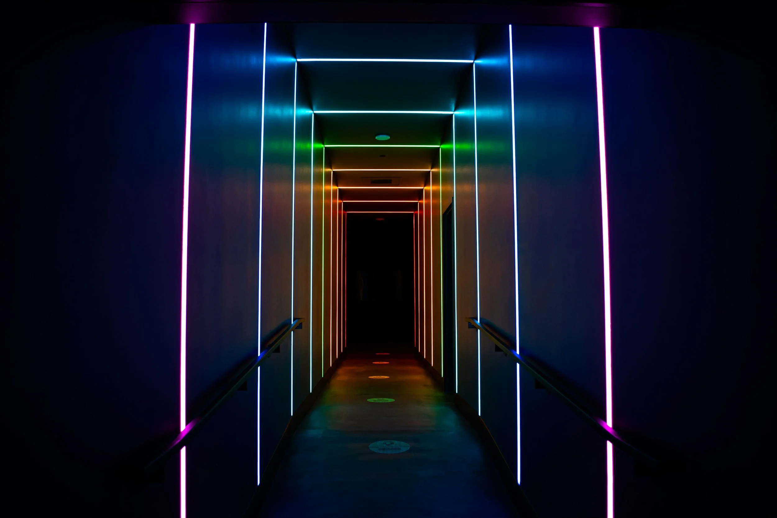 Dark hallway illuminated with colorful neon lights forming vertical and horizontal lines, creating a tunnel effect.