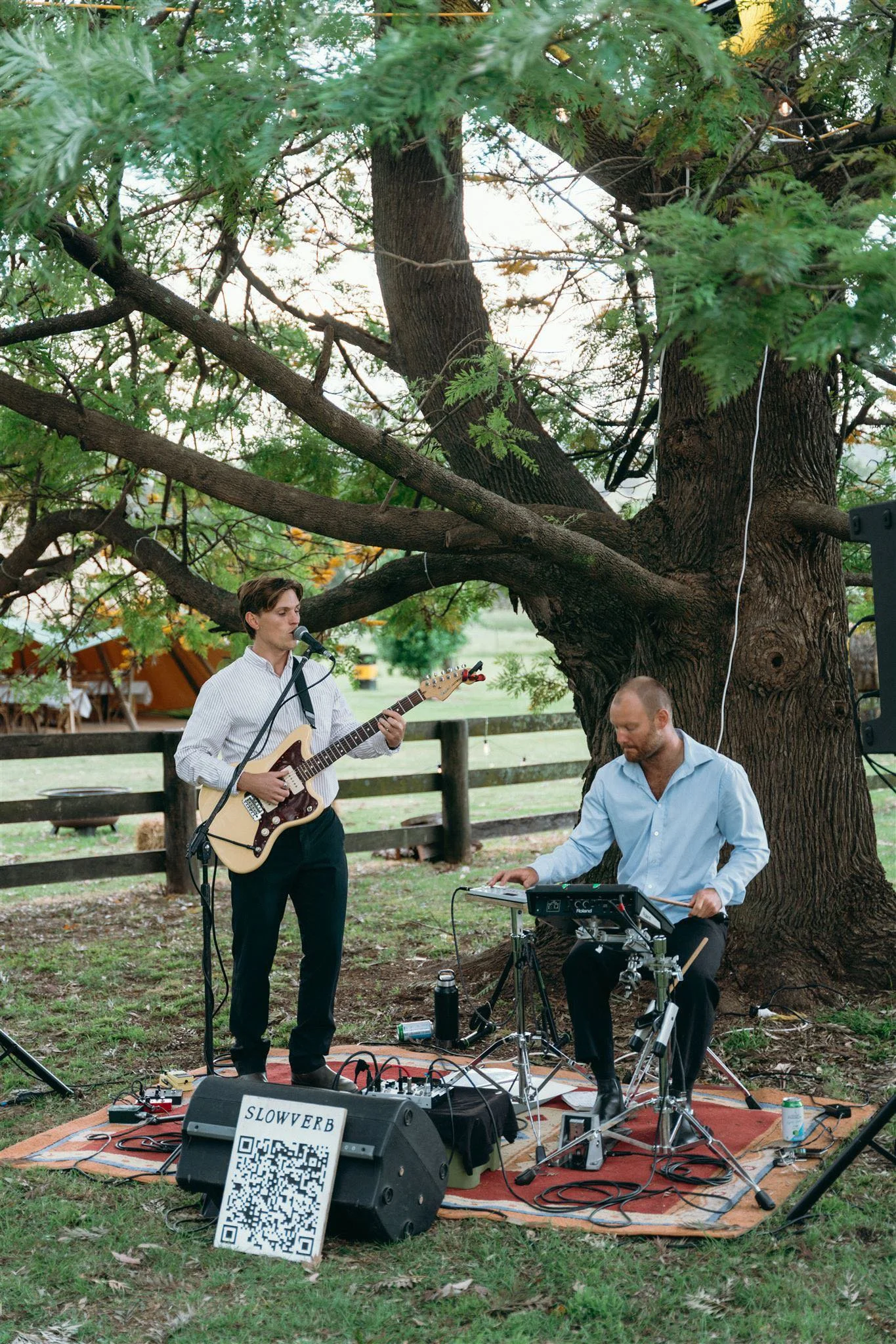 Slowverb wedding duo performing live acoustic music outdoors beneath a tree, featuring guitar and electronic percussion.