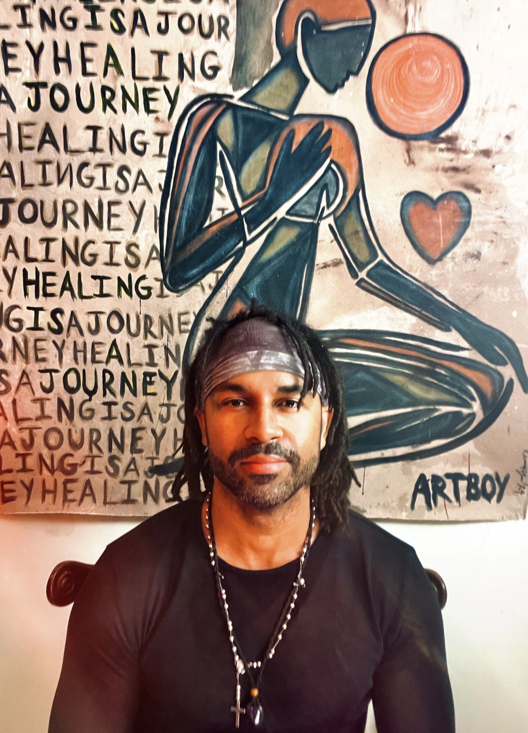 A man with dreadlocks wearing a black shirt, a gray headband, and a necklace with a cross, is sitting in front of a colorful abstract painting of a meditating figure with one hand on their chest and attentive to a glowing orb and a heart symbol, with the words 'Healing is a journey' repeatedly written in the background.