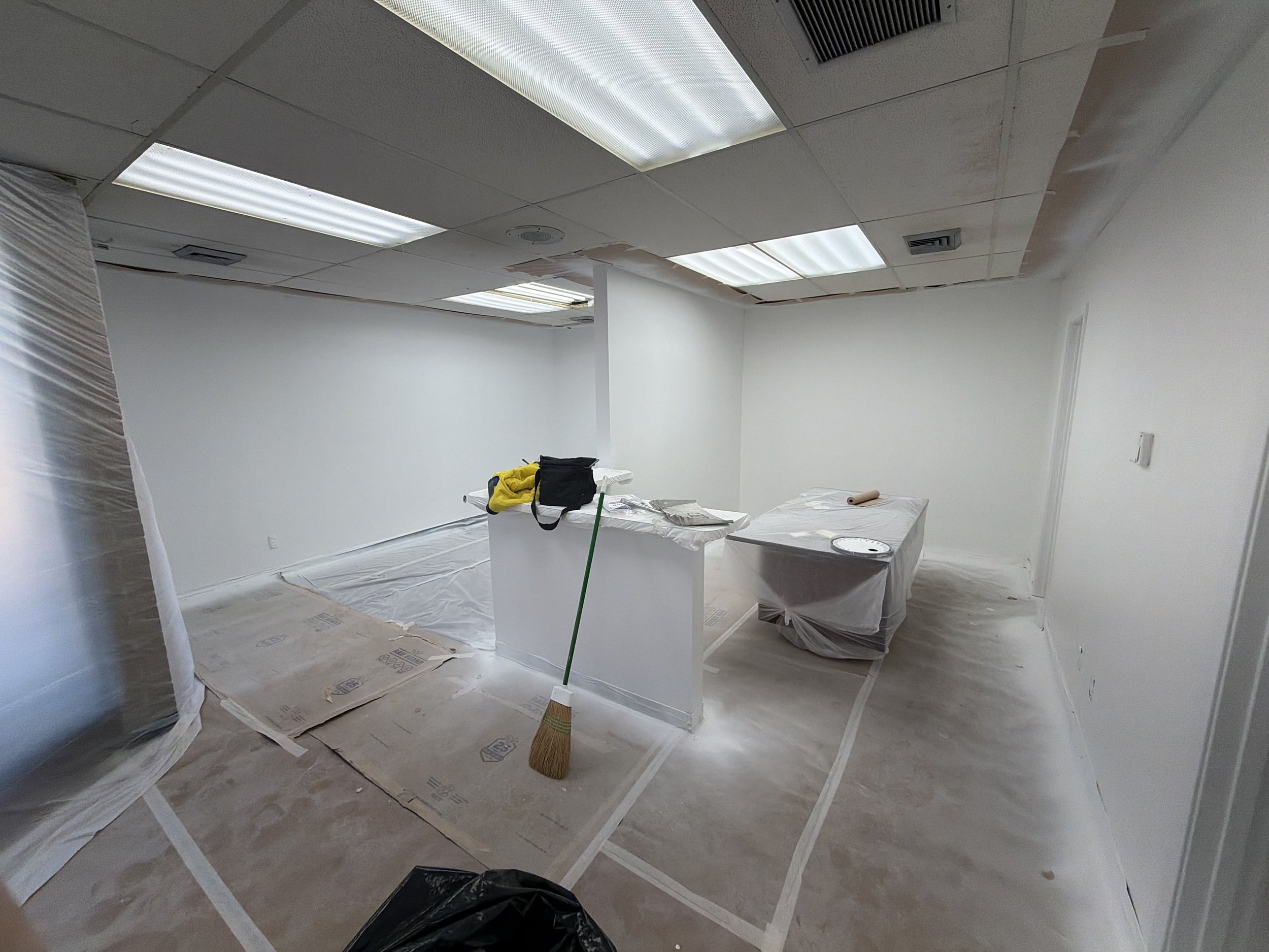 An office space under renovation with white walls and ceiling, protected with plastic and paper coverings. There is a broom leaning against a small white counter, and the ceiling tiles and light fixtures are visible, some of which are damaged or miss
