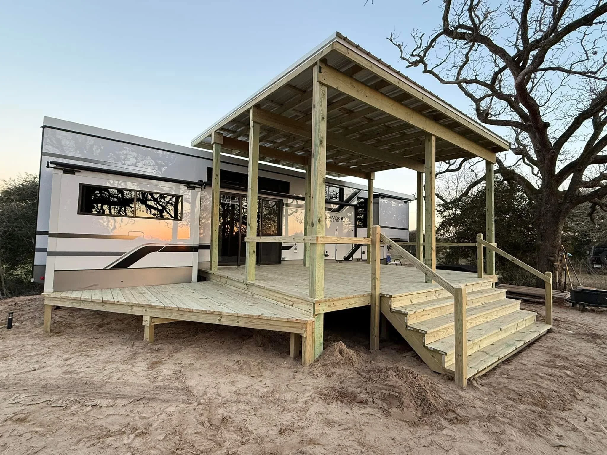 A newly built wooden deck with stairs and a covered patio area in front of a camper trailer on bare ground, with large leafless trees in the background at sunset.