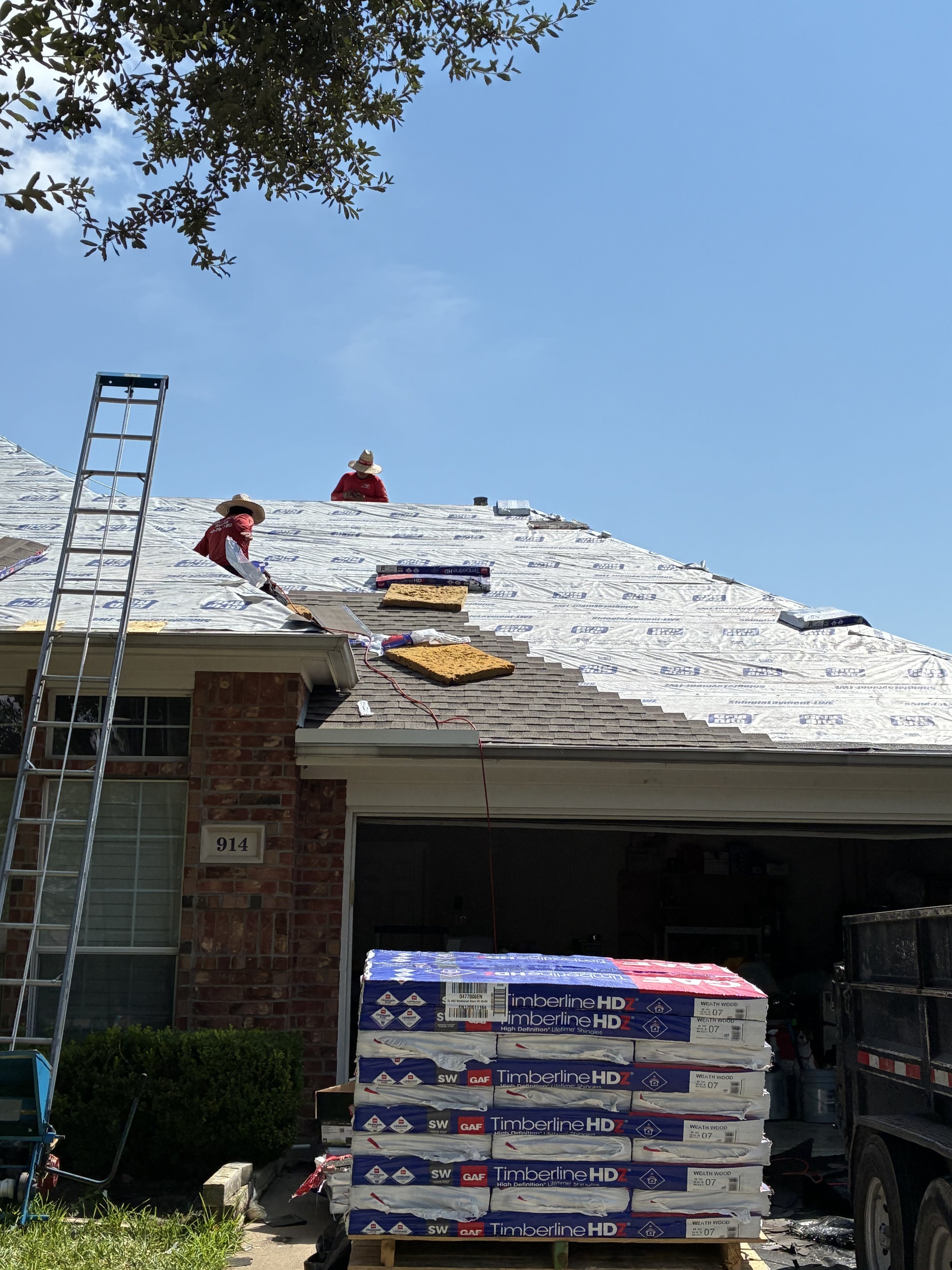 Workers roofing a house with shingles, with new shingles stacked in front, under a clear blue sky.