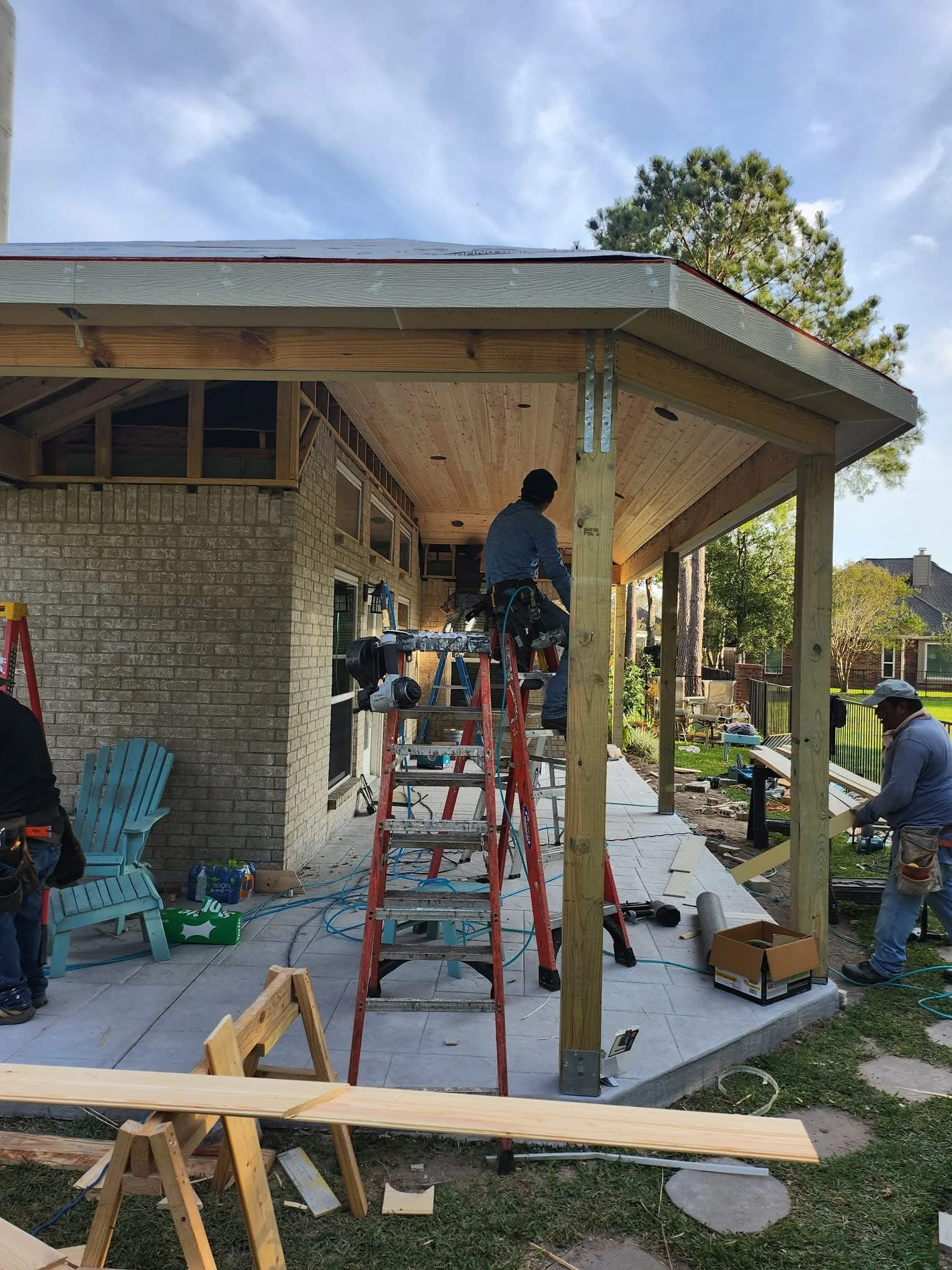 Construction workers building a porch or extension on a house with wooden beams and a tiled patio, surrounded by tools and construction materials.