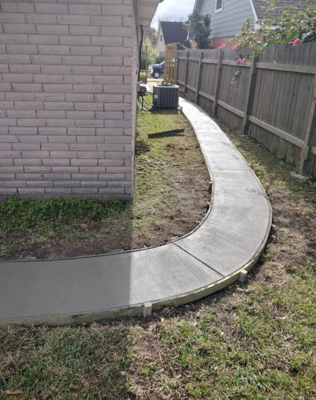 Curved concrete sidewalk next to a brick house, with a wooden fence on the right and a grassy yard on the left.