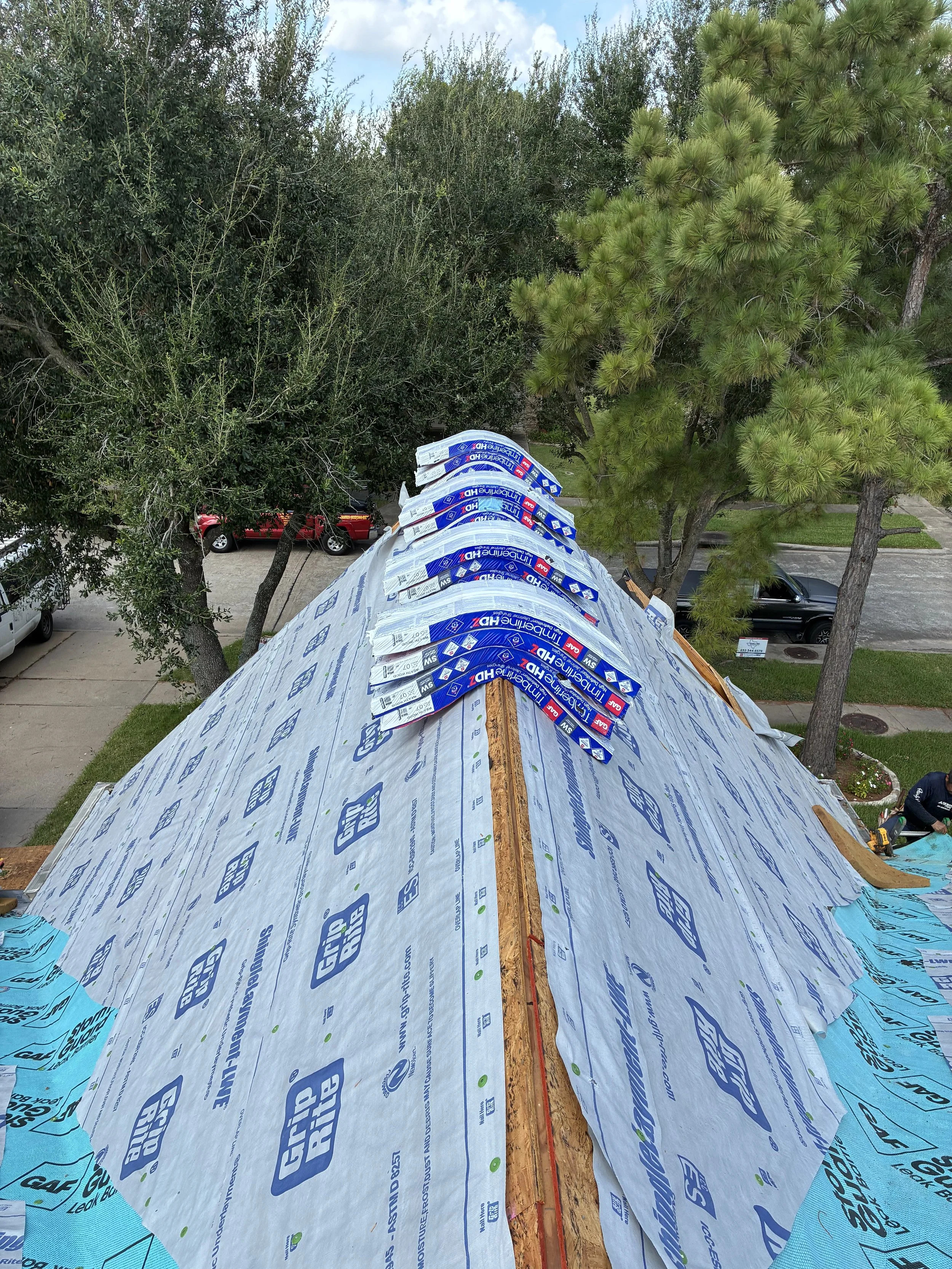 View from the roof of a building under construction, showing roofing materials and tools, with trees and parked cars in the background.