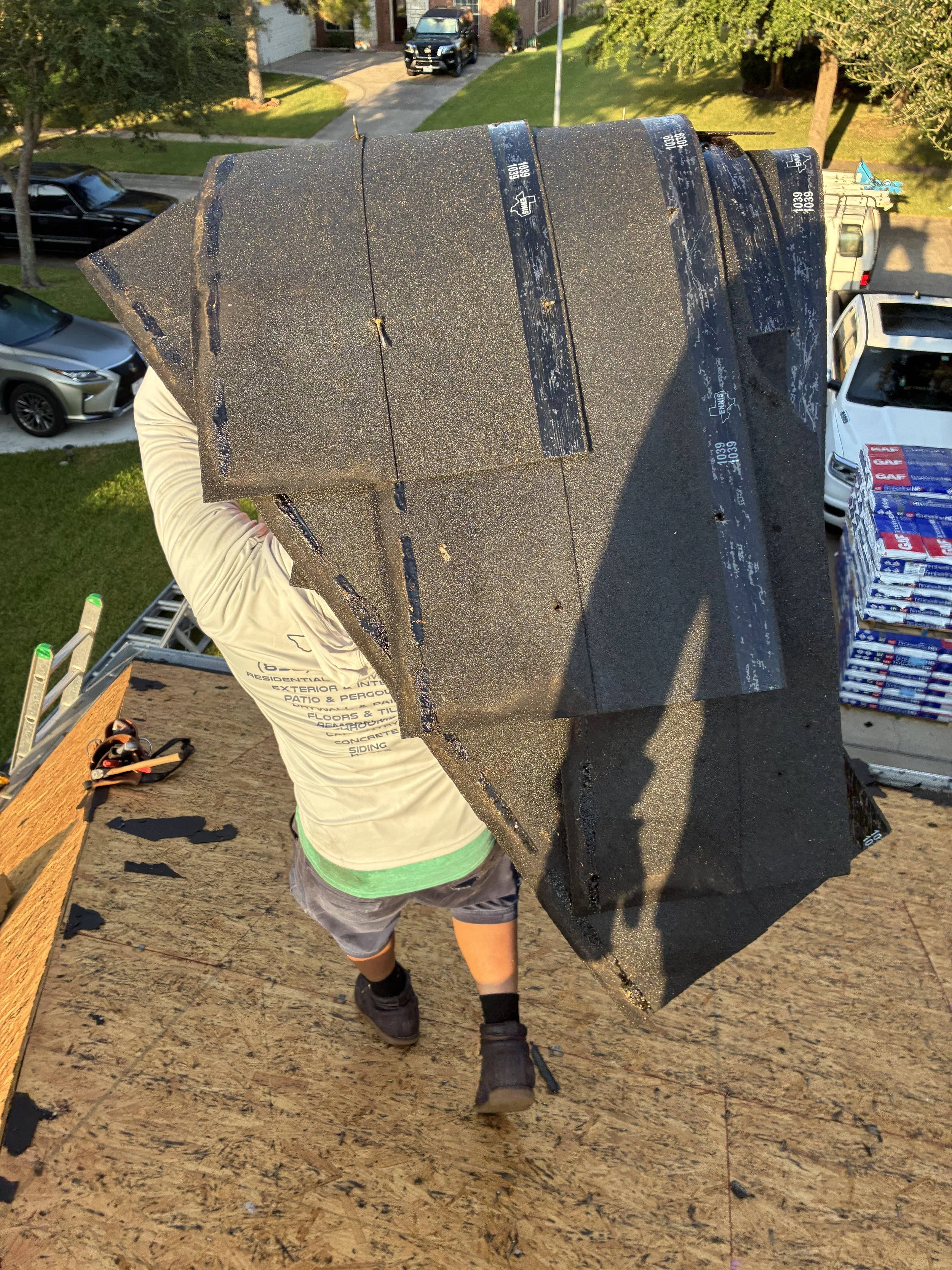 A person working on a rooftop, holding a bundle of roofing shingles over their shoulder, with a sprained ankle visible in a supportive brace. The roof is covered with black shingles, and in the background, there are trees, a driveway, and parked cars