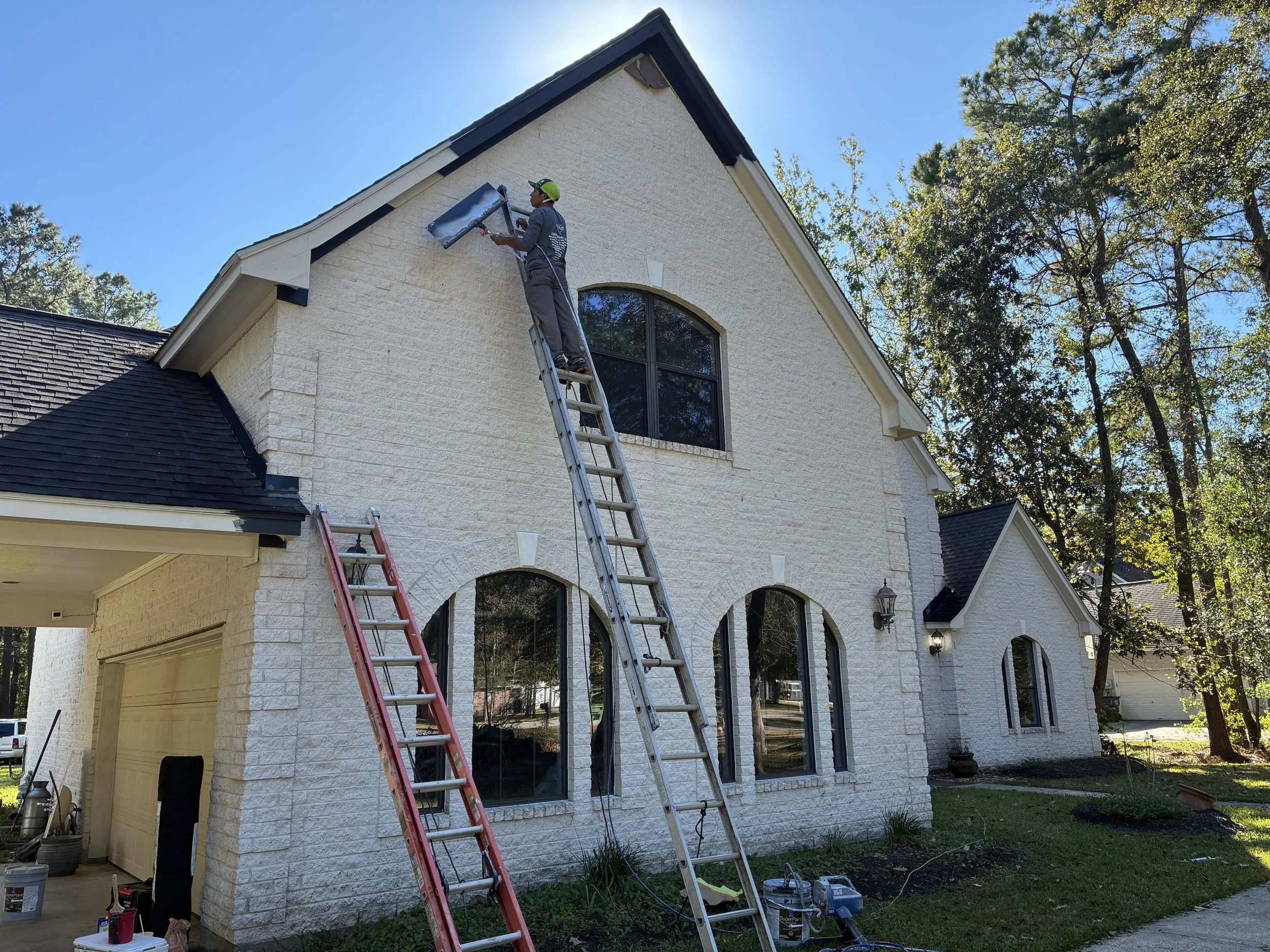 Person working on the exterior of a white brick house, standing on a ladder, installing or repairing a gutter, surrounded by trees and tools.