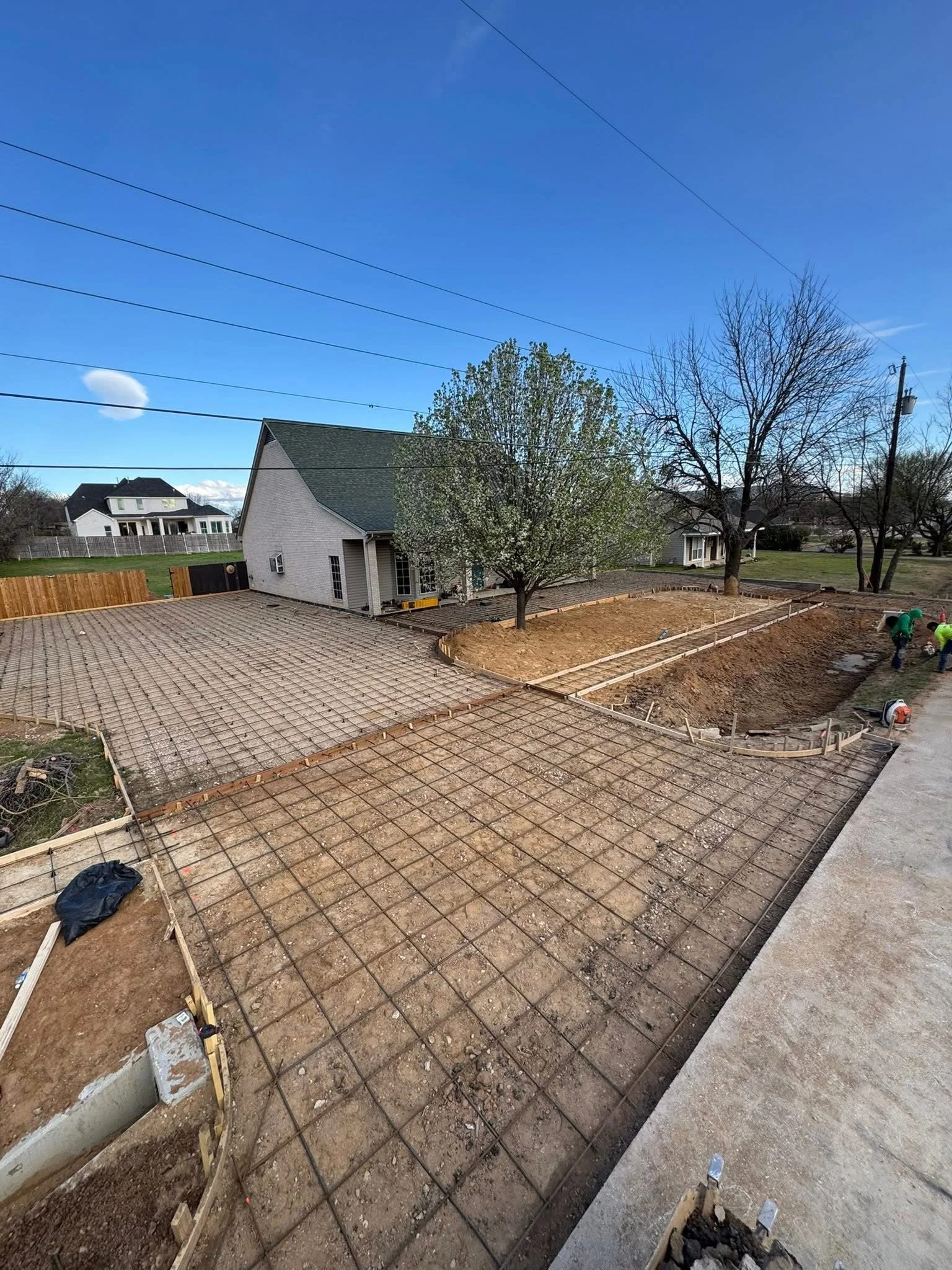 Construction site with rebar grid for concrete pouring in a backyard, next to a house with a green roof and blooming trees.