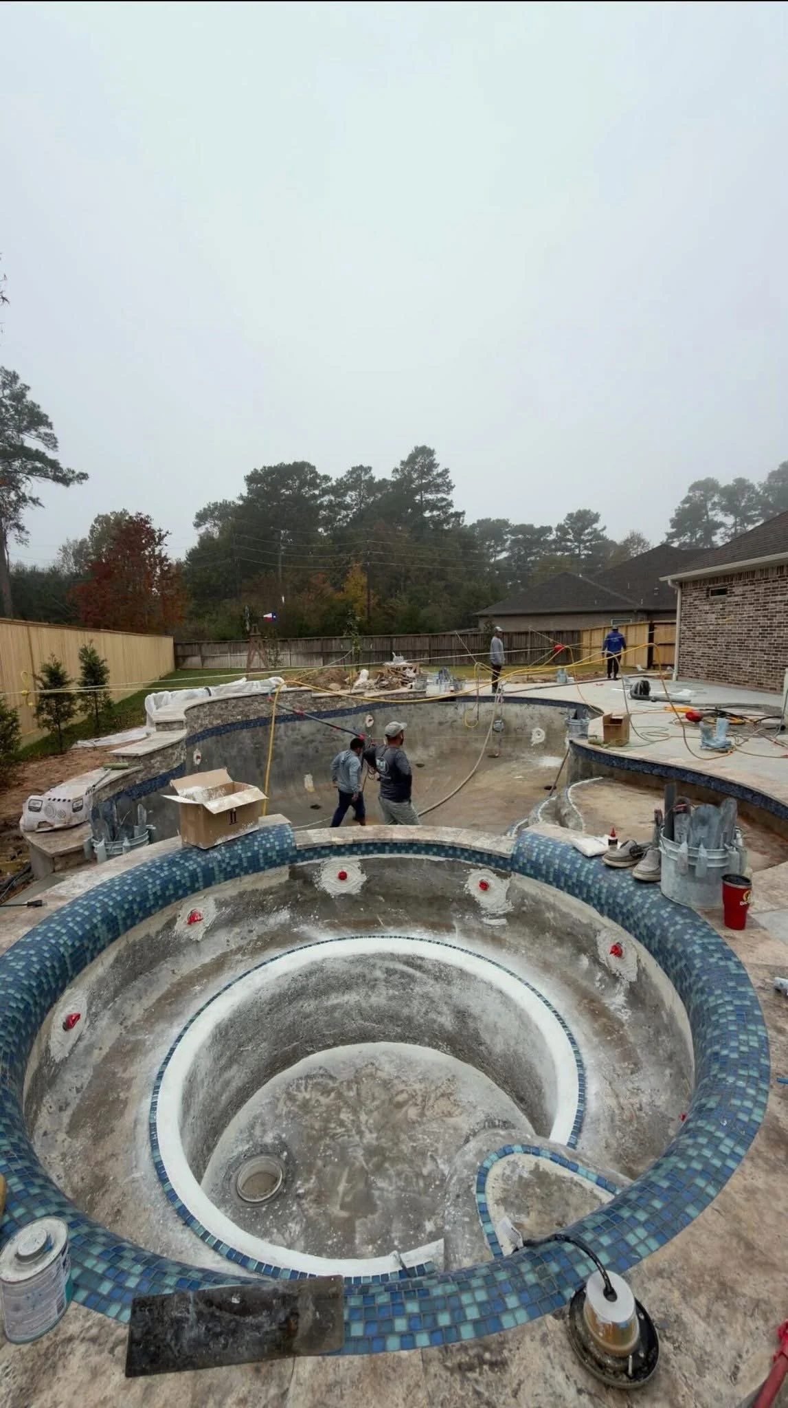 Under construction of a backyard pool with a hot tub, with workers present, surrounded by tools and construction materials, on a foggy day.