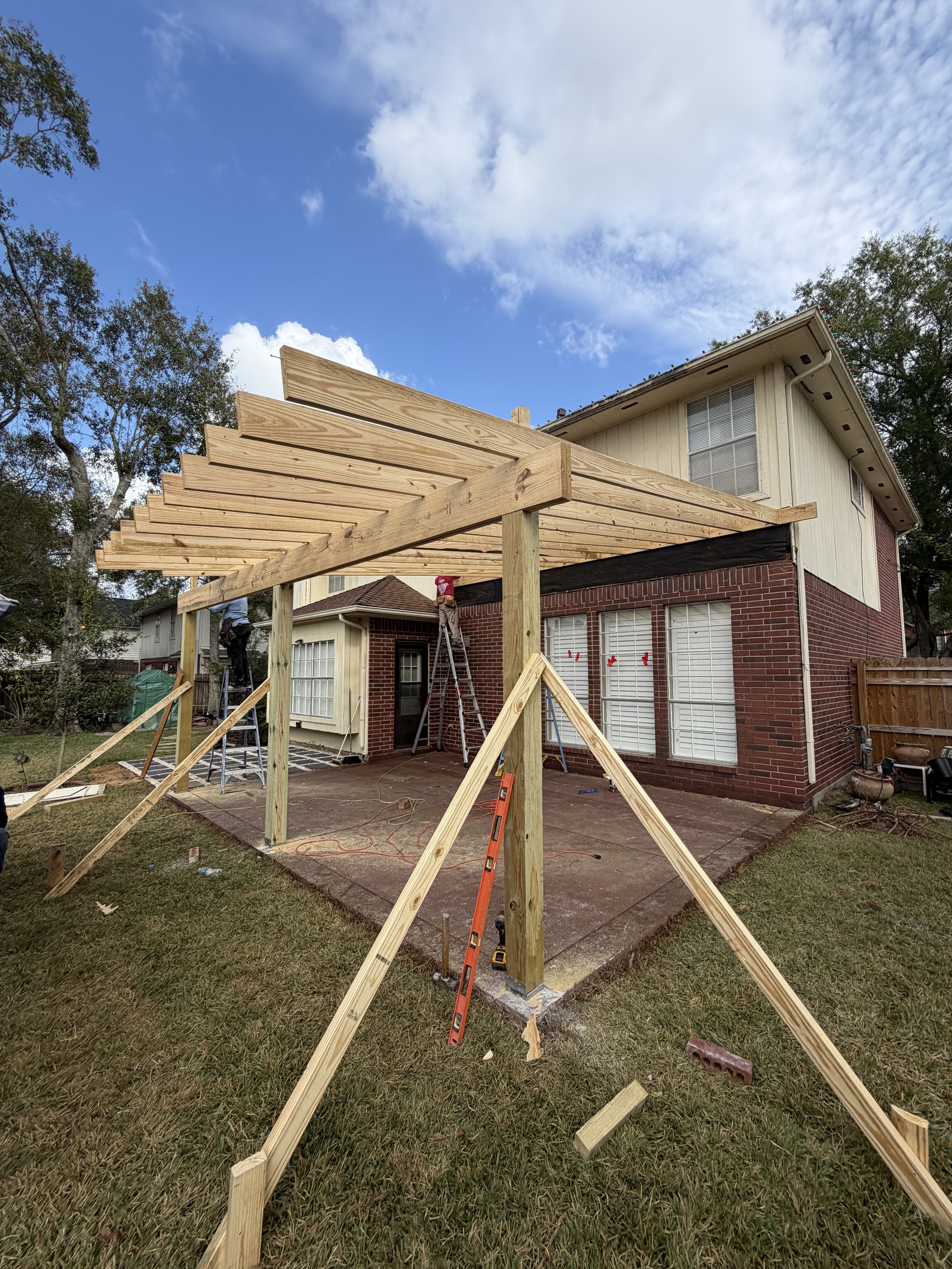 Construction of a wooden pergola structure attached to a backyard house, with workers on ladders, on a partly cloudy day.