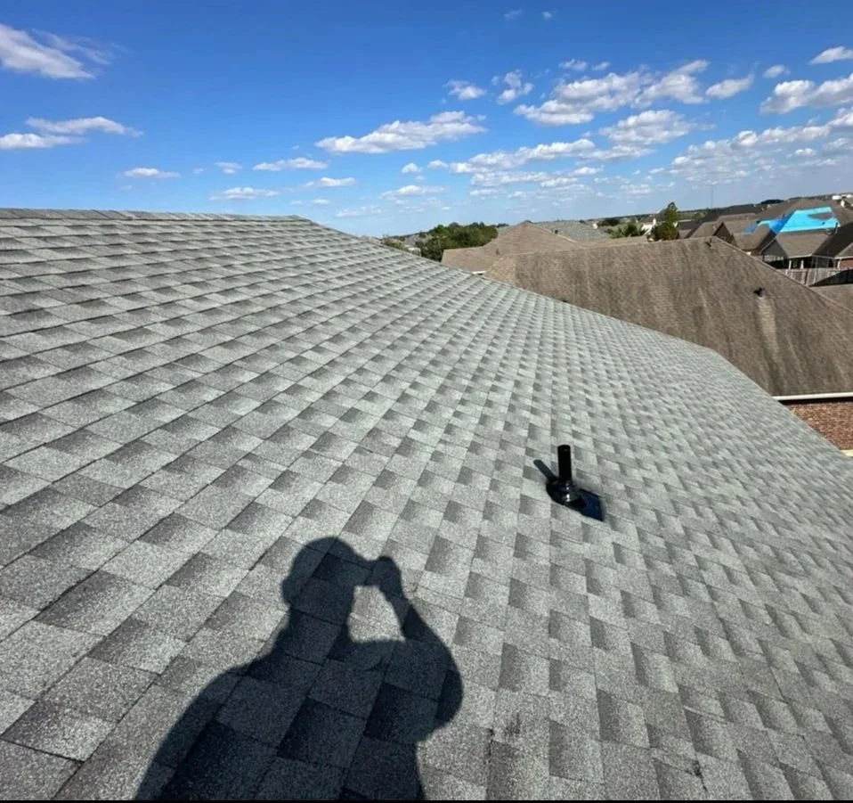 View of a residential rooftop with gray asphalt shingles, under a partly cloudy blue sky, with neighboring houses visible in the distance.