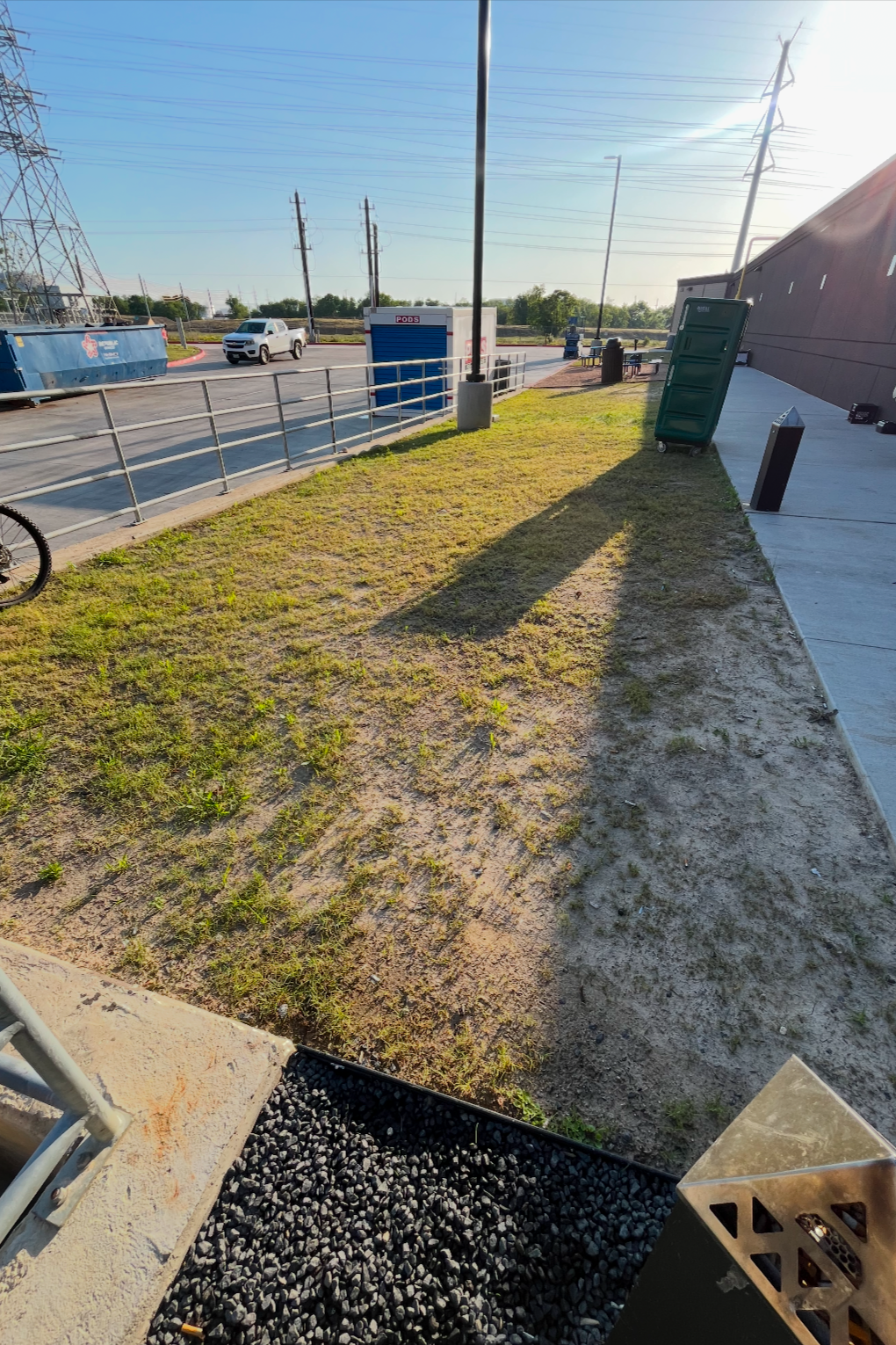 A small patch of grass and dirt surrounded by a sidewalk and a street with cars. In the background, there are power lines, utility poles, and a small blue building with a sign reading 'PODS'. A green trash bin is tipped over on the sidewalk, and the 