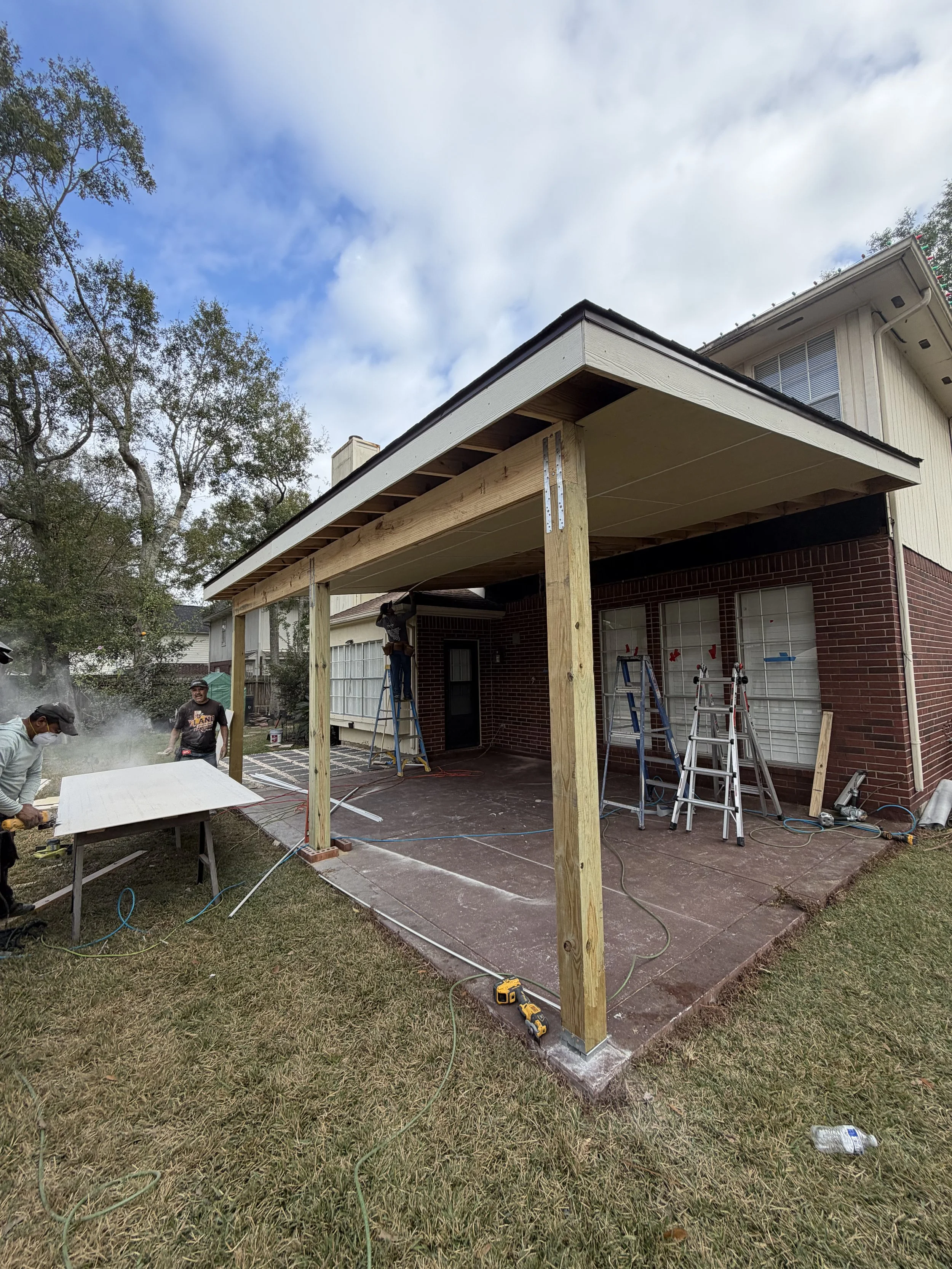Construction workers building a covered patio or porch extension on a brick house, with ladders, tools, and wooden beams.