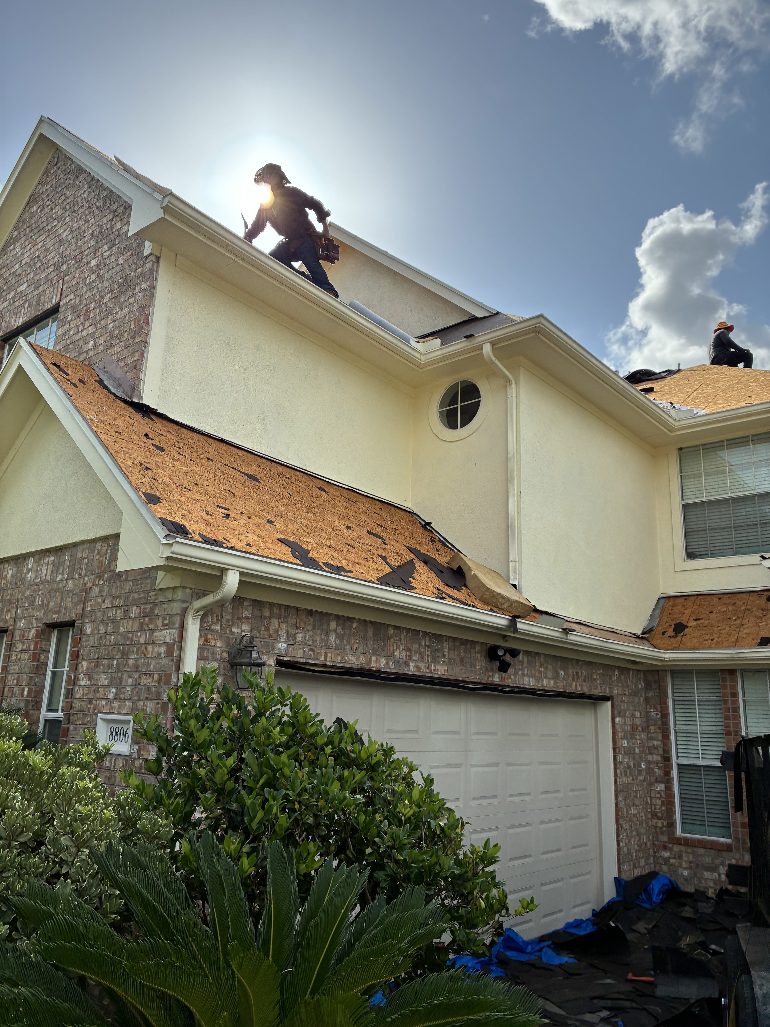 Construction workers repairing the roof of a two-story house with orange shingles, some of which are damaged or missing. The house has a brick facade on the lower level and light-colored siding on the upper level. There is a bush in the foreground an