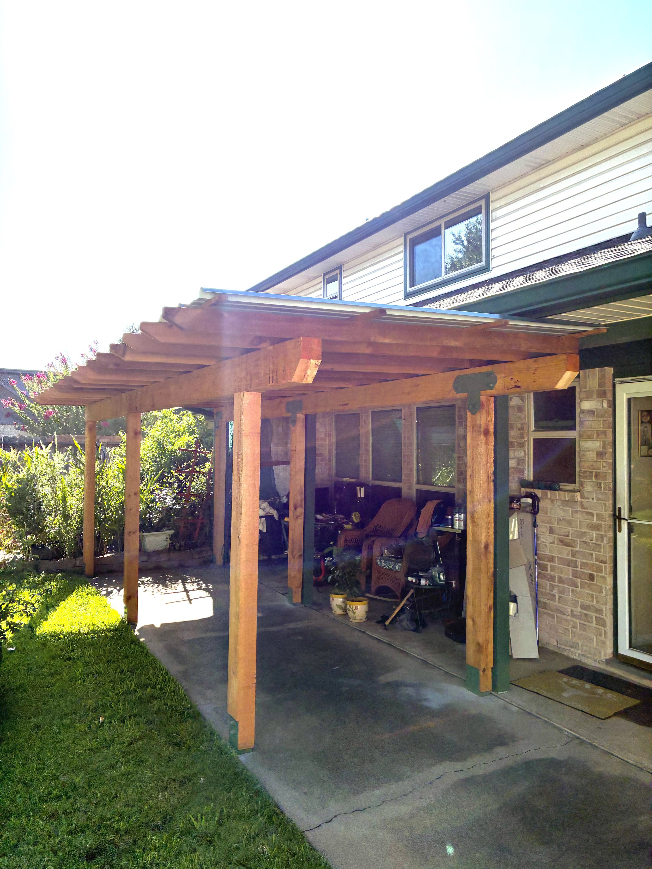 A residential house with a wooden pergola structure in the backyard, featuring garden chairs and gardening supplies, sunny weather, and lush greenery nearby.