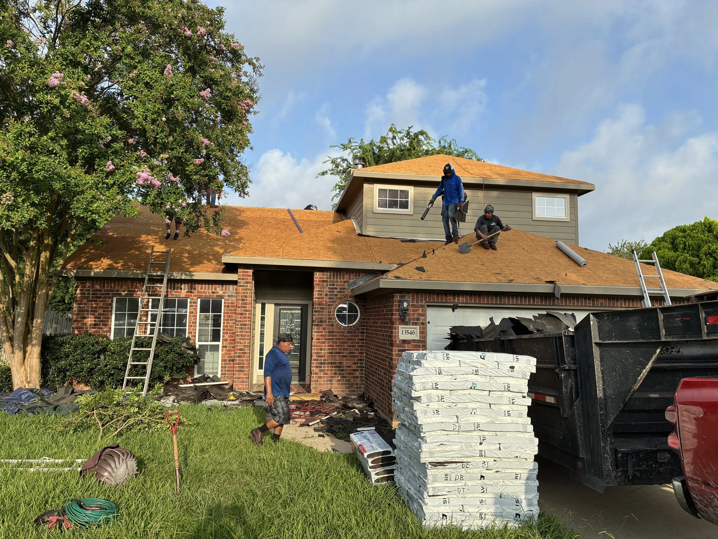 Workers replacing the roof of a brick house with shingles. Some workers are on the roof, while one is on the ground. There is a ladder leaning against the house, and a truck parked nearby filled with roofing materials.