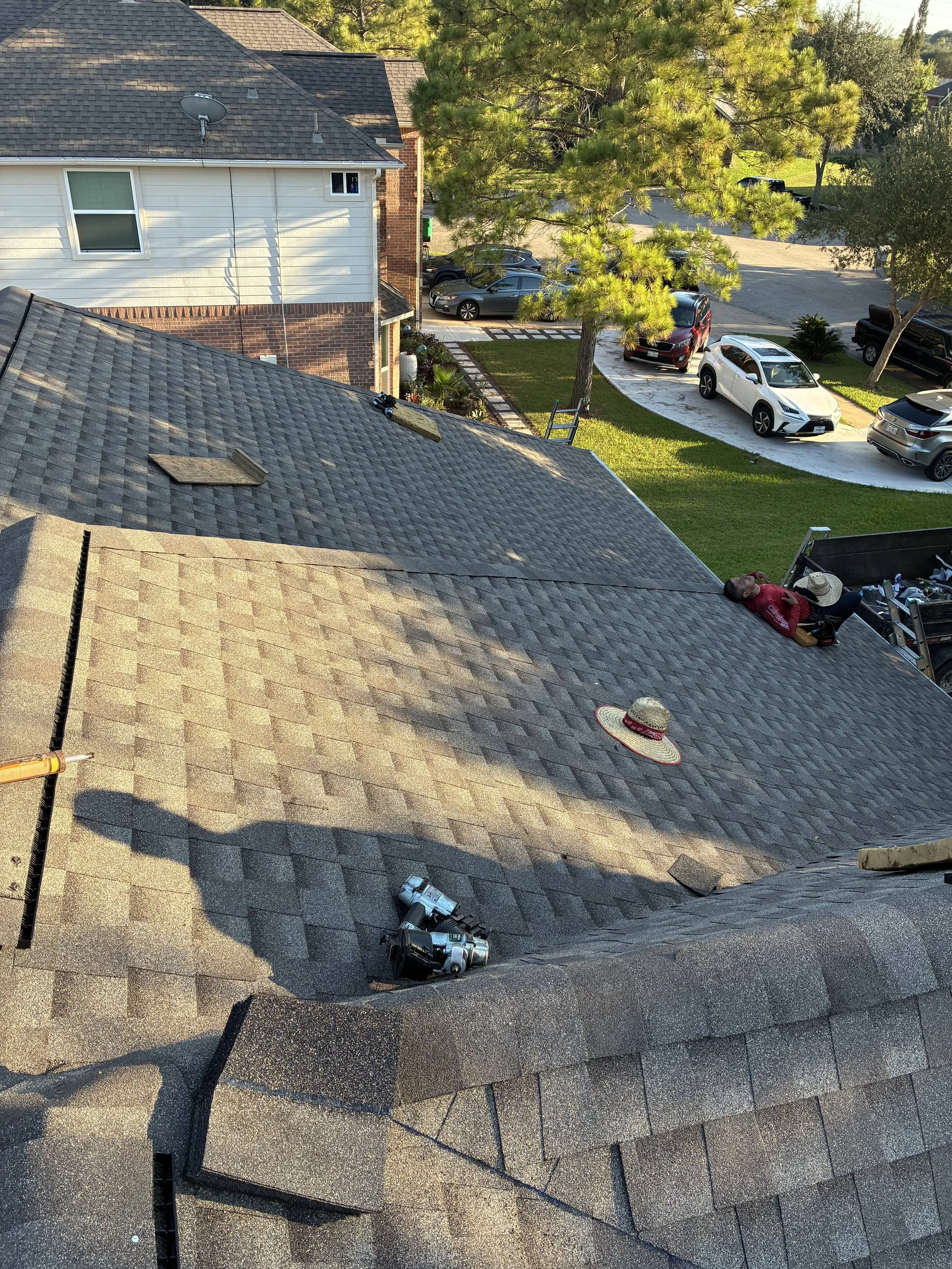 View of a residential roof undergoing maintenance with a worker lying on the roof, tools scattered, and neighboring houses, trees, and a parking lot with cars in the background.