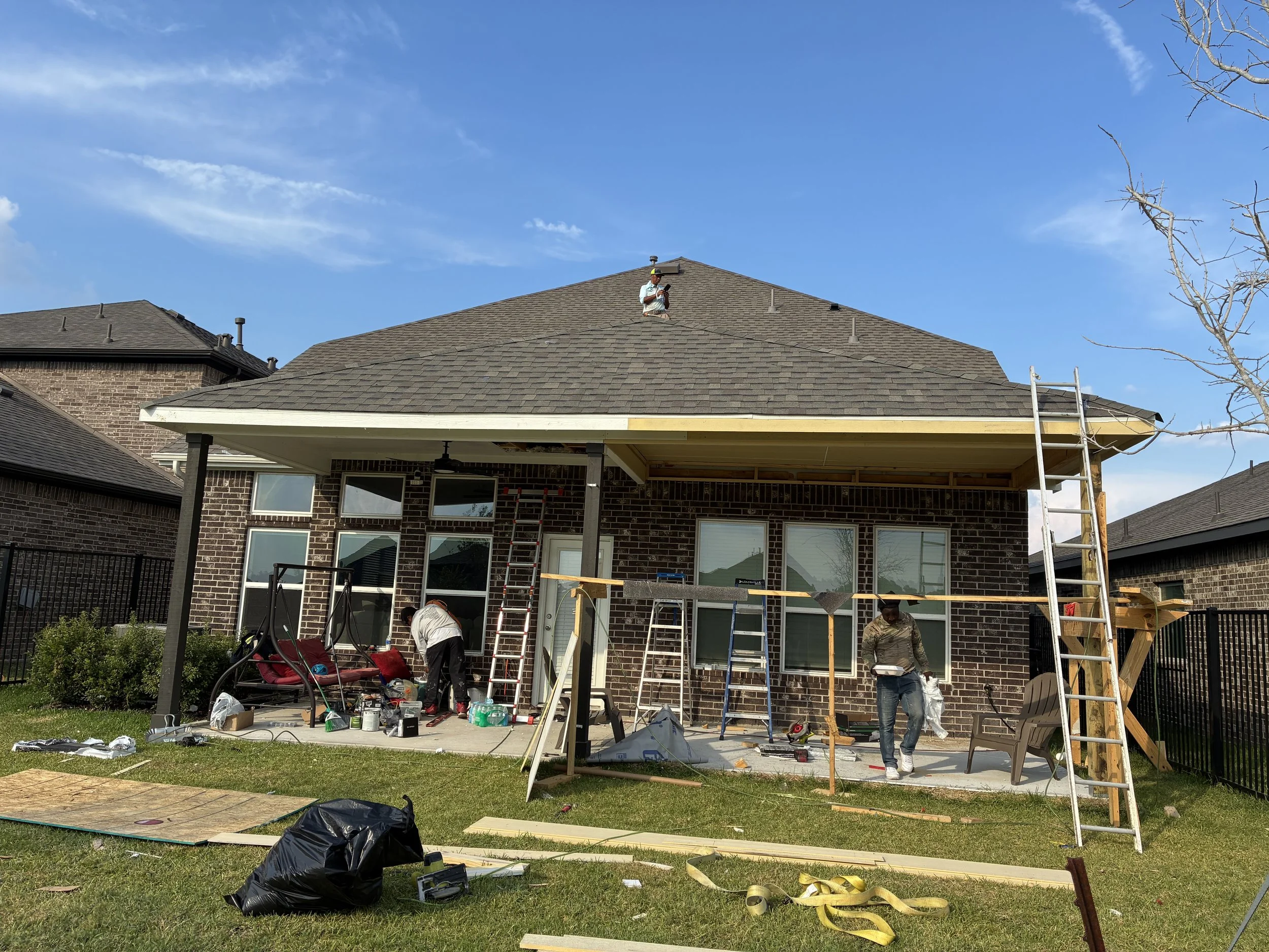 Construction workers building a new porch or patio area at the back of a brick house, with scaffolding and tools around.