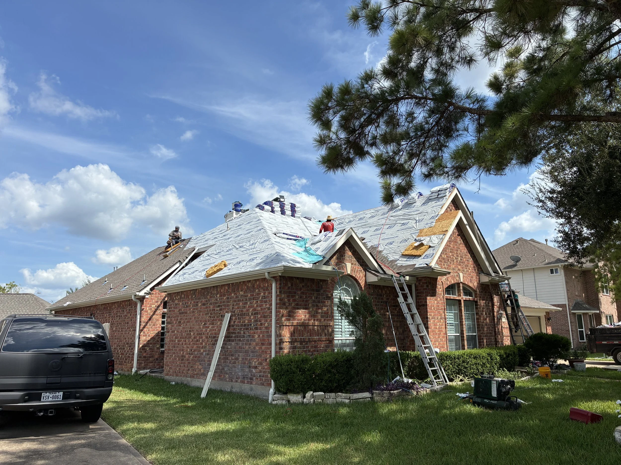 A brick house with a steep roof under construction or renovation, with workers on the roof working on new shingles, ladders leaning against the house, and construction tools on the ground. The sky is partly cloudy, and there is a tree in the foregrou