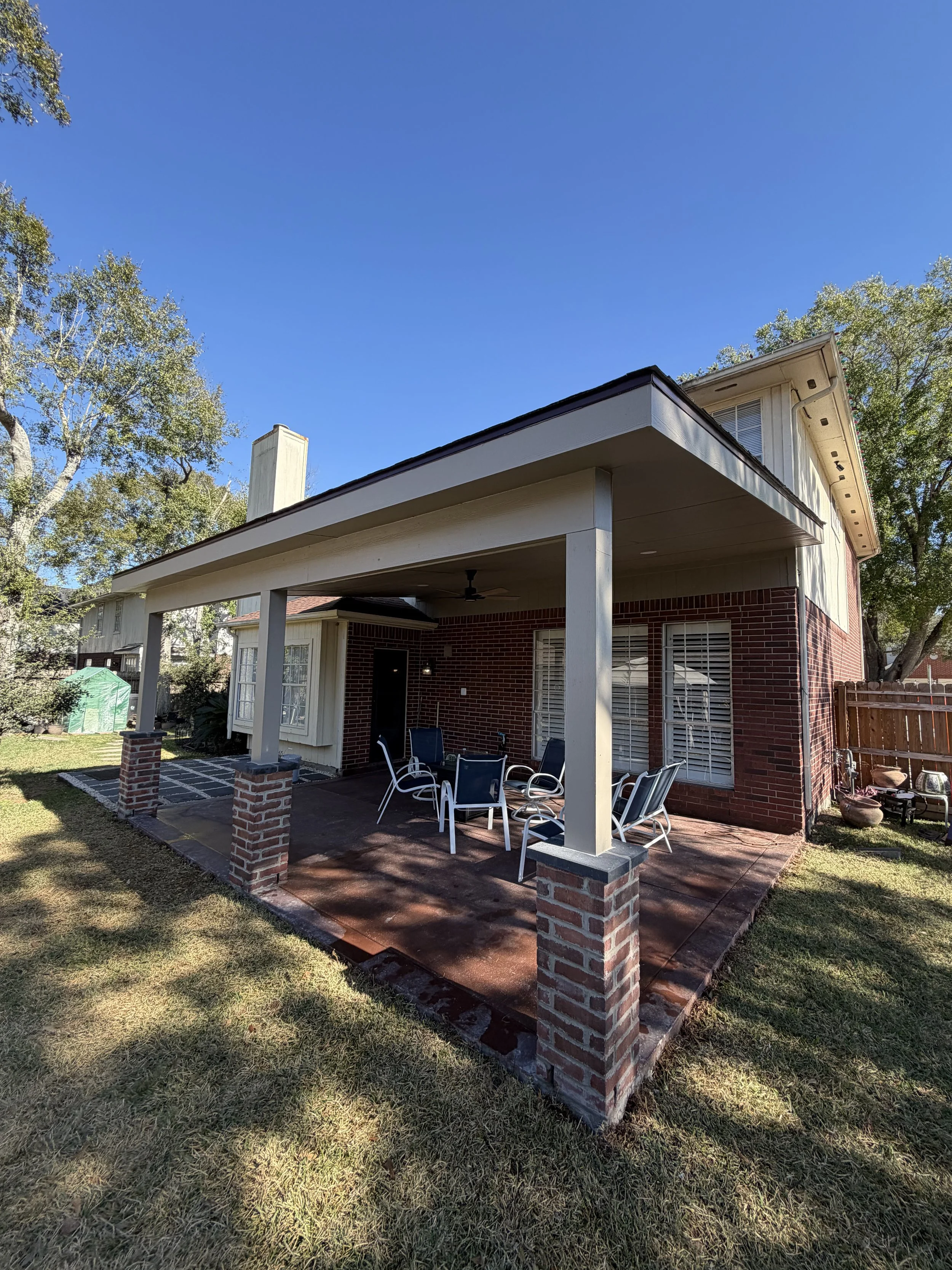 Backyard patio with a covered seating area, brick pillars, outdoor chairs, and a wooden fence, with trees and clear blue sky in the background.