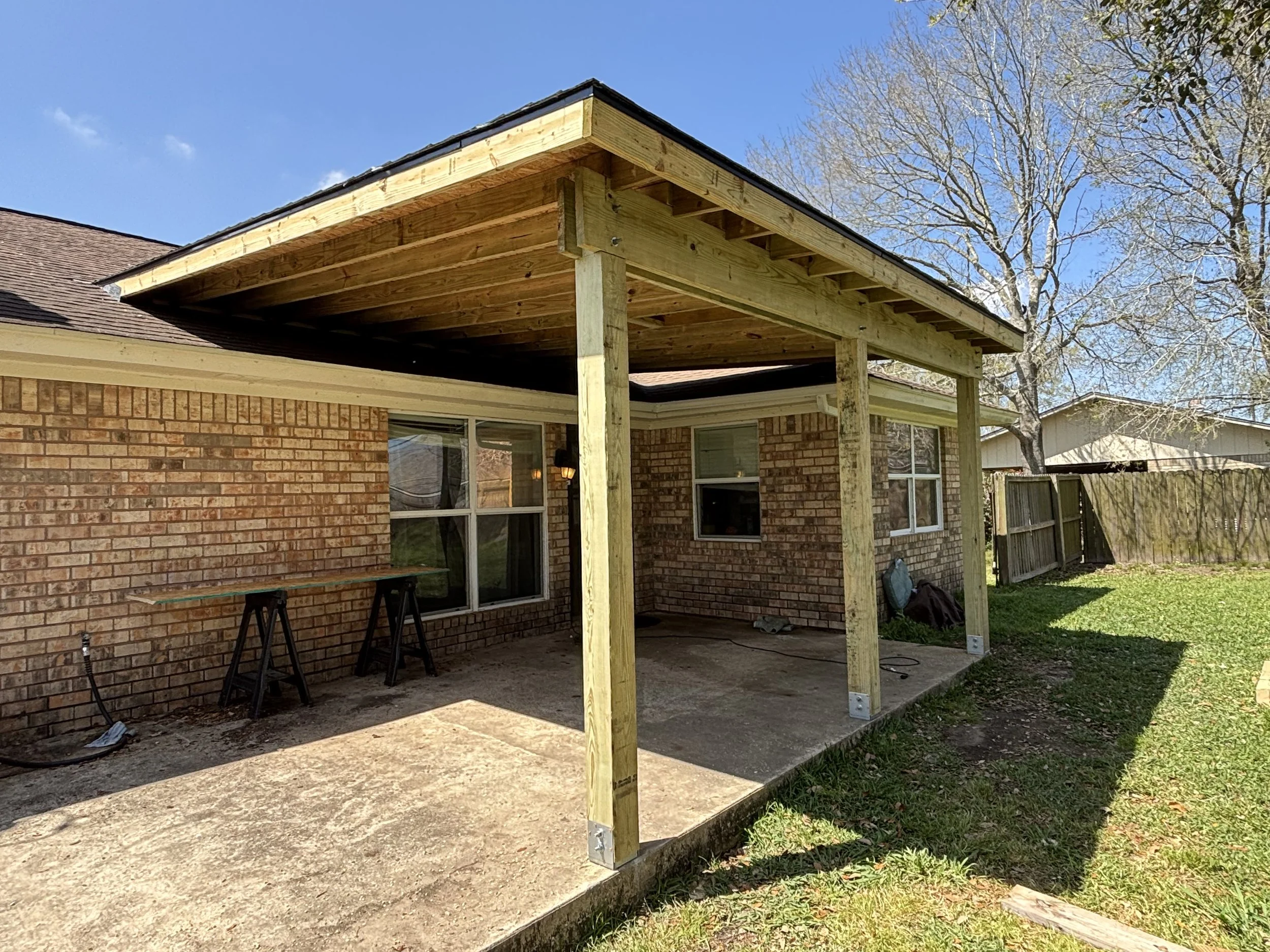 Backyard area with brick house, under-construction wooden patio cover, and a concrete slab, with some tools and equipment leaning against the house.