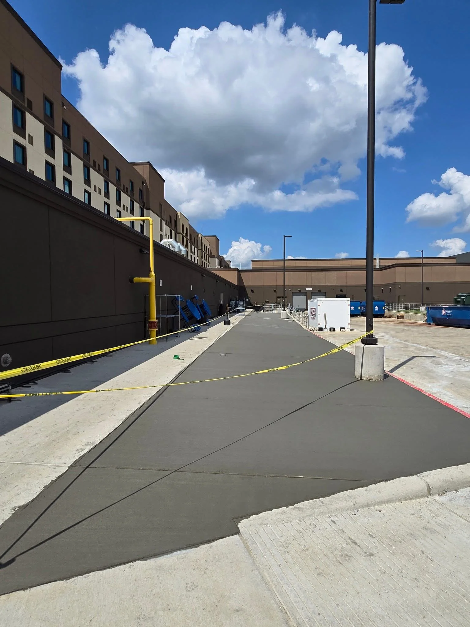 Sidewalk under construction with yellow caution tape, blue recycling bins, and a brown building with multiple windows in the background on a partly cloudy day.
