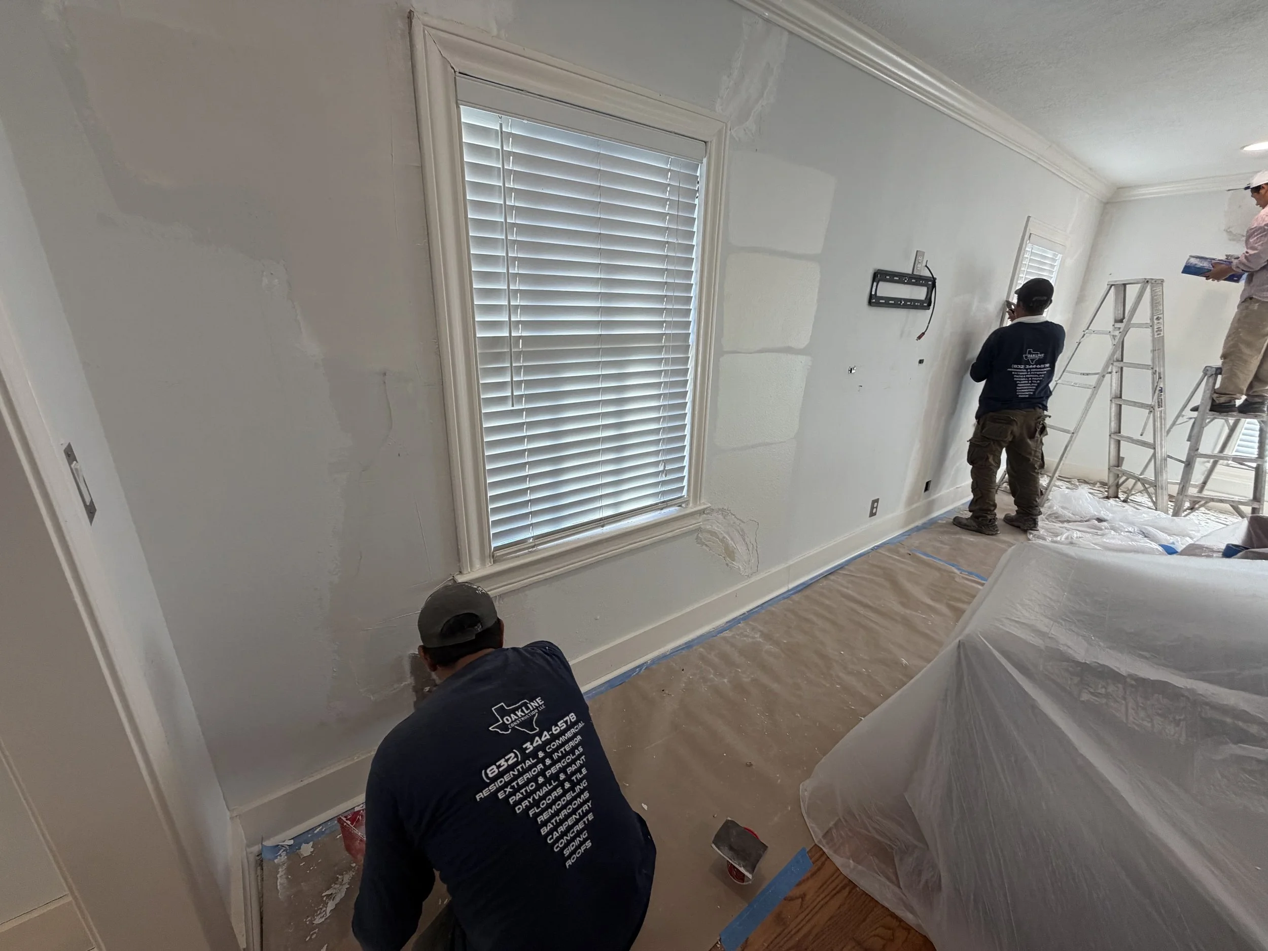 Workers applying paint or primer to the walls in a room under renovation, with ladders and covered furniture.